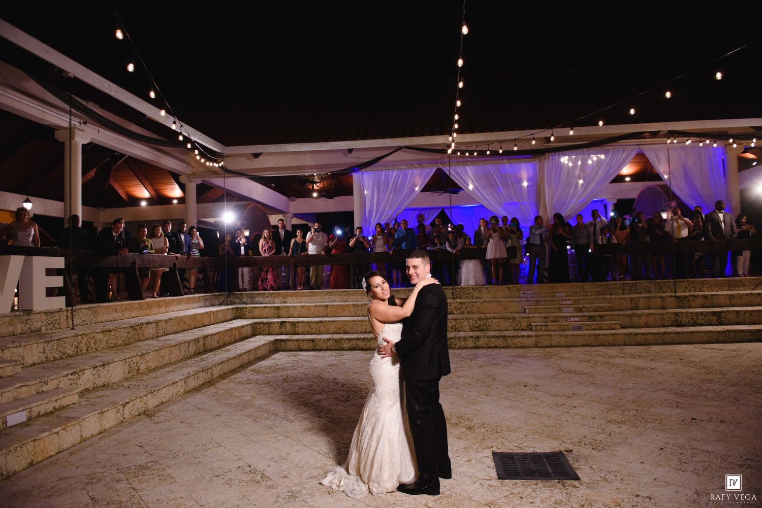 Bride and groom share first dance outdoors at elegant night wedding reception with guests watching under string lights.