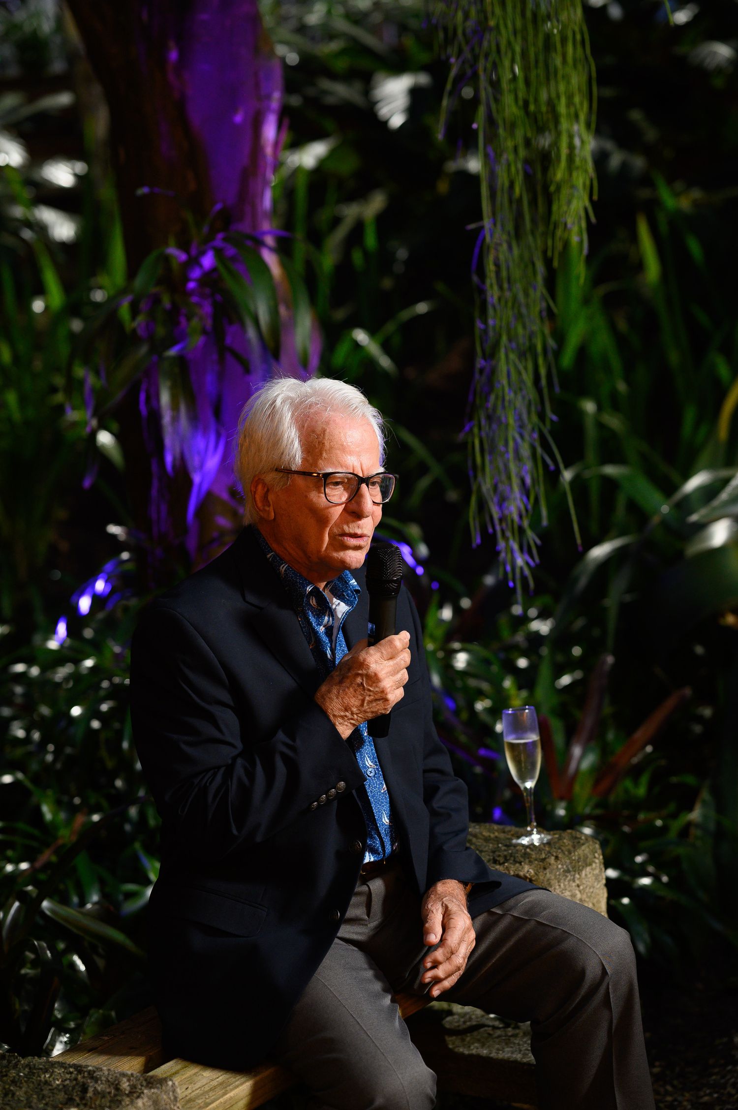 Elderly man with white hair and glasses speaking into a microphone while seated outdoors among lush green plants.