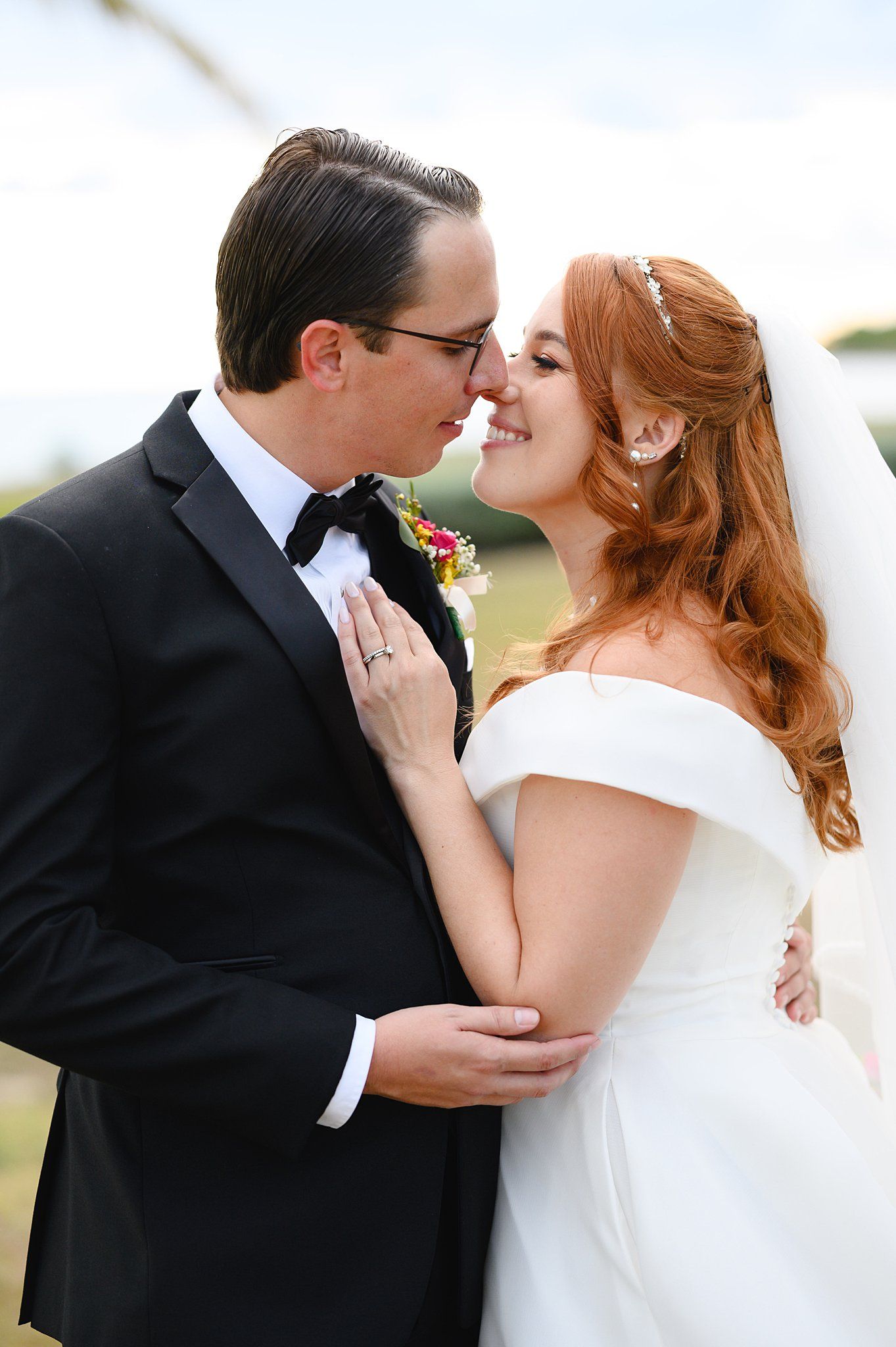 Bride and groom sharing a romantic moment, she in white off-shoulder gown, he in black tuxedo, outdoors.