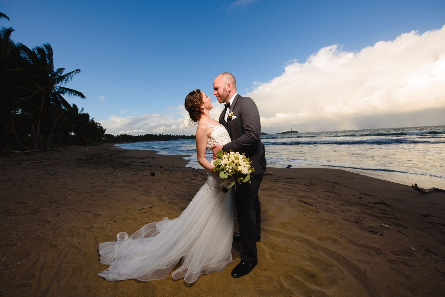 Bride and groom share a romantic moment on a tropical beach at sunset, with palm trees and ocean waves in the background.