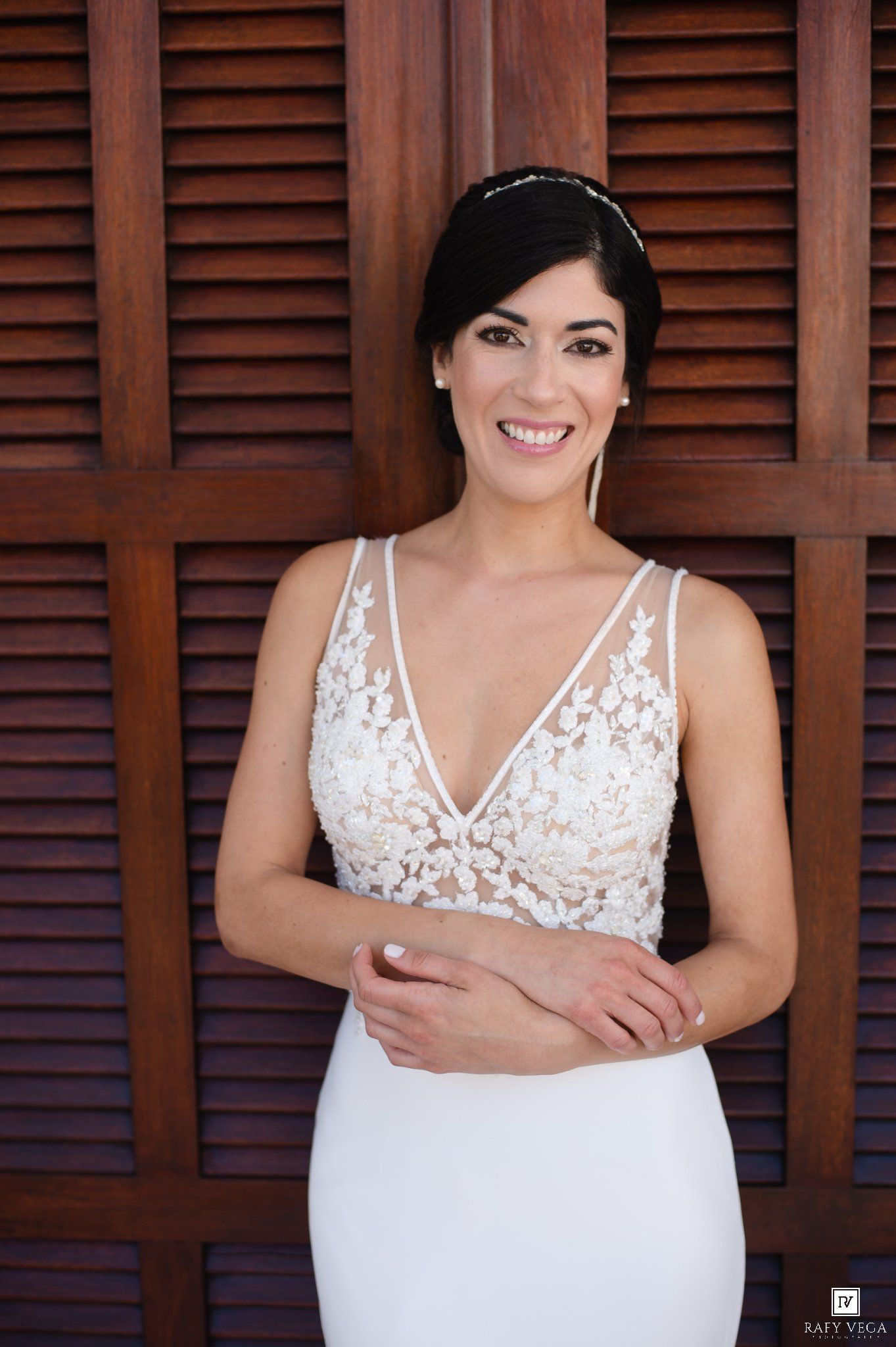Smiling bride in a white lace V-neck wedding gown posing against wooden shutters backdrop.