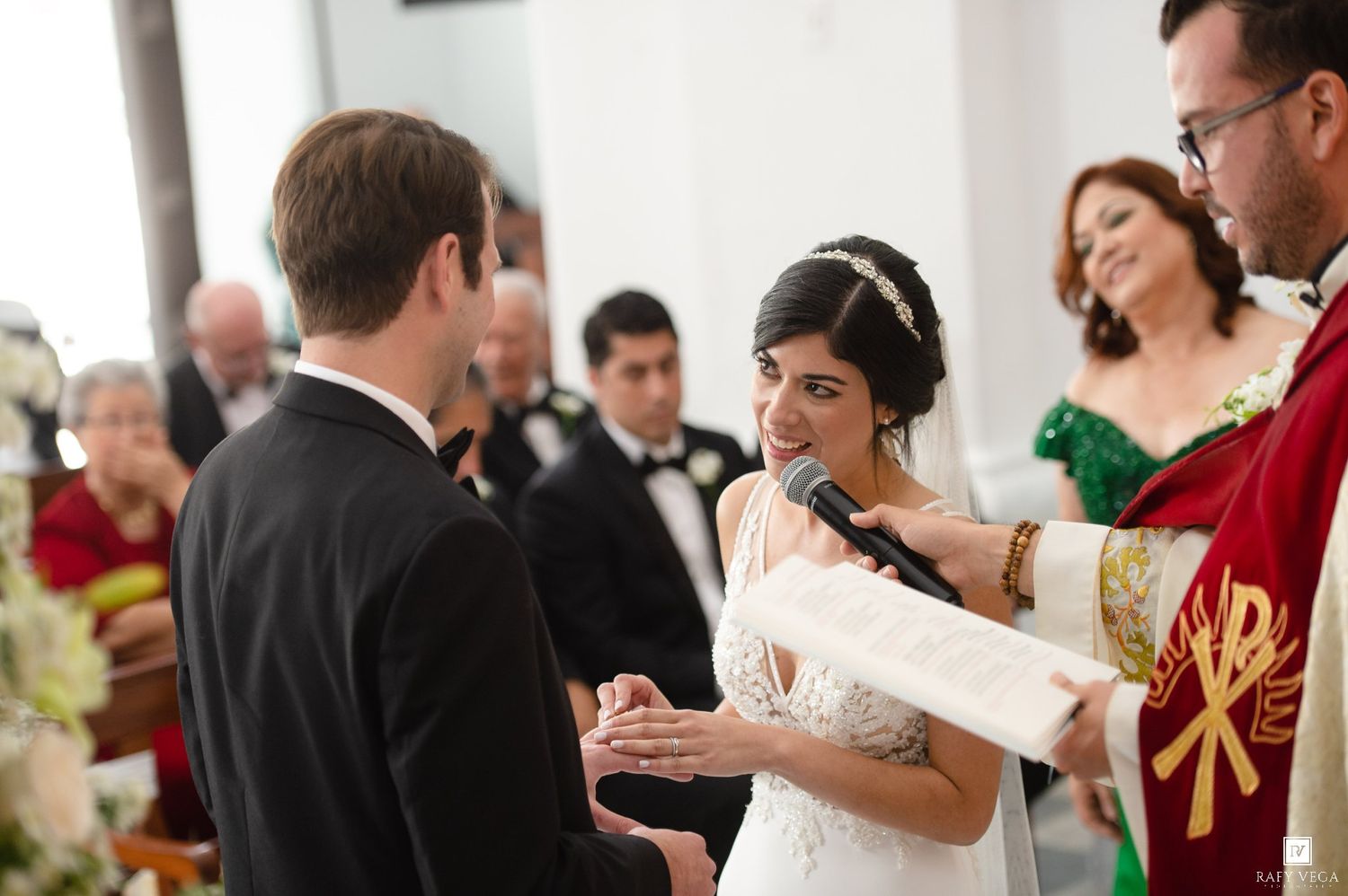 Bride and groom exchange vows during elegant wedding ceremony, officiant holding microphone in decorated church.