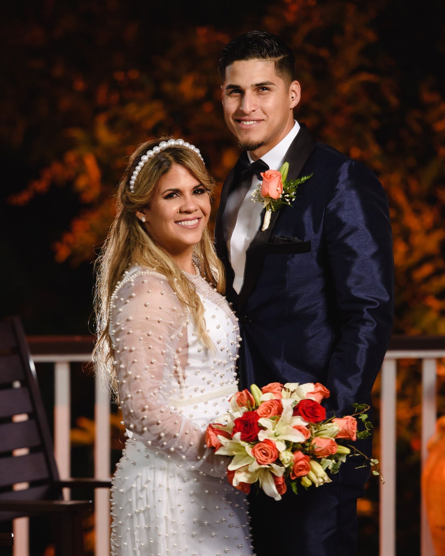 Smiling bride in white pearl dress holding coral bouquet poses with groom in navy tuxedo at outdoor wedding.