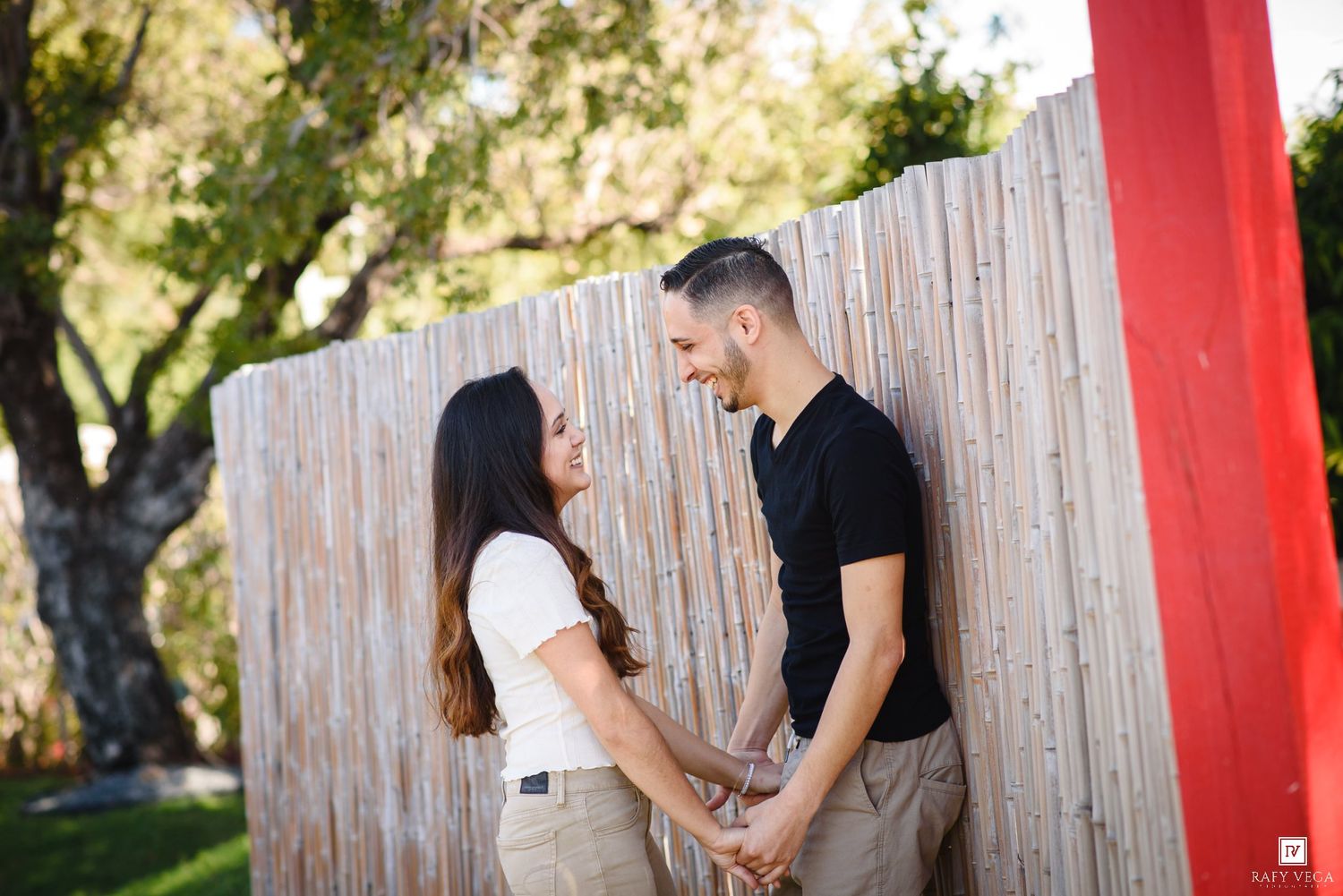 A couple holds hands and smiles at each other beside a wooden fence outdoors.