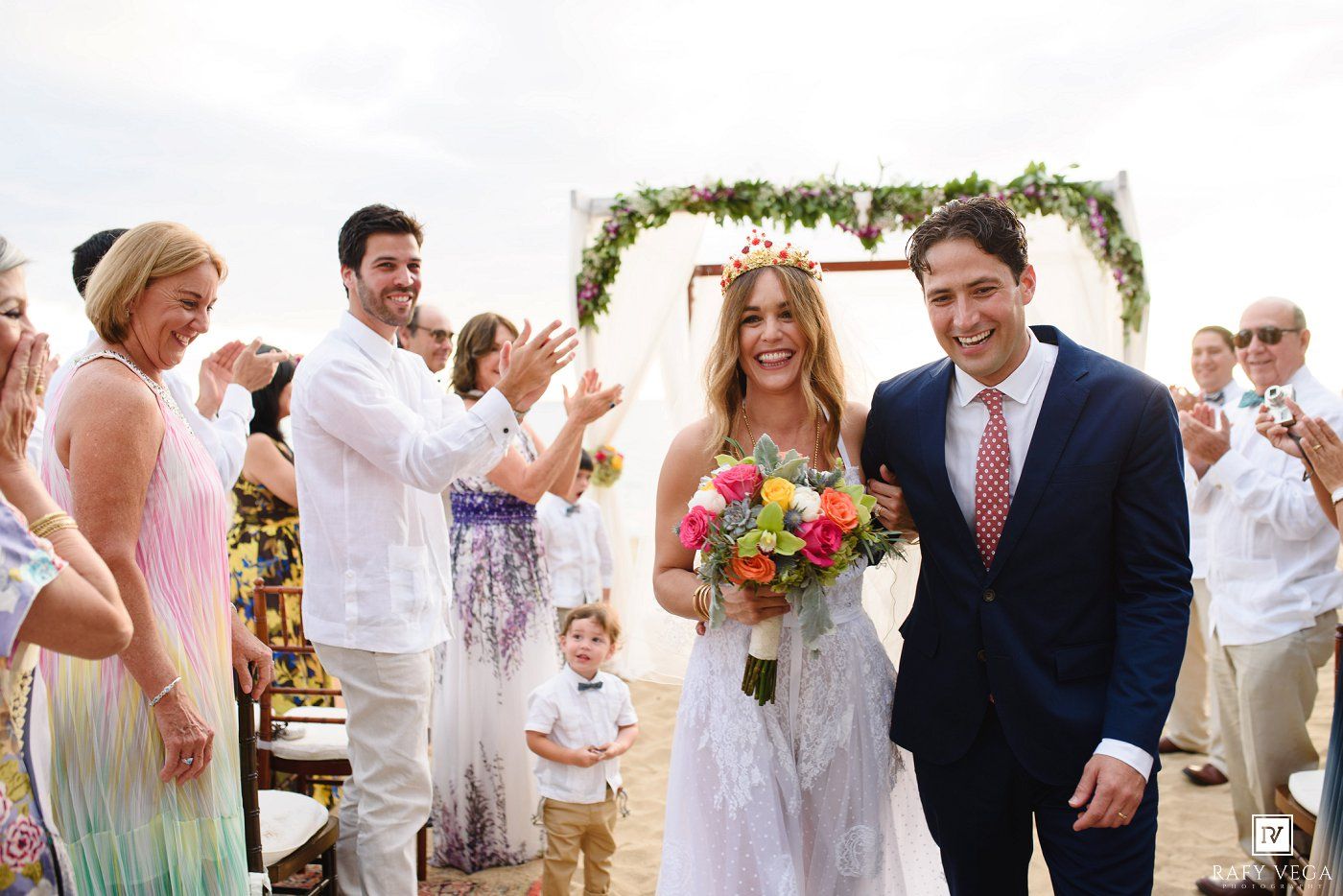 Bride and groom walk down beach wedding aisle, guests applaud, floral arch backdrop, bride holds colorful bouquet.