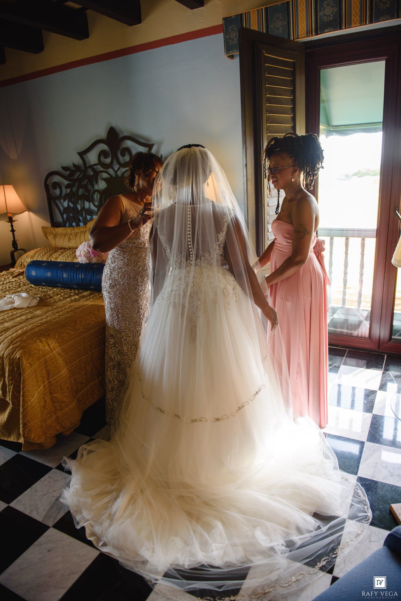 A bride in a lace gown with veil is assisted by two women in a elegant room with checkered floors.