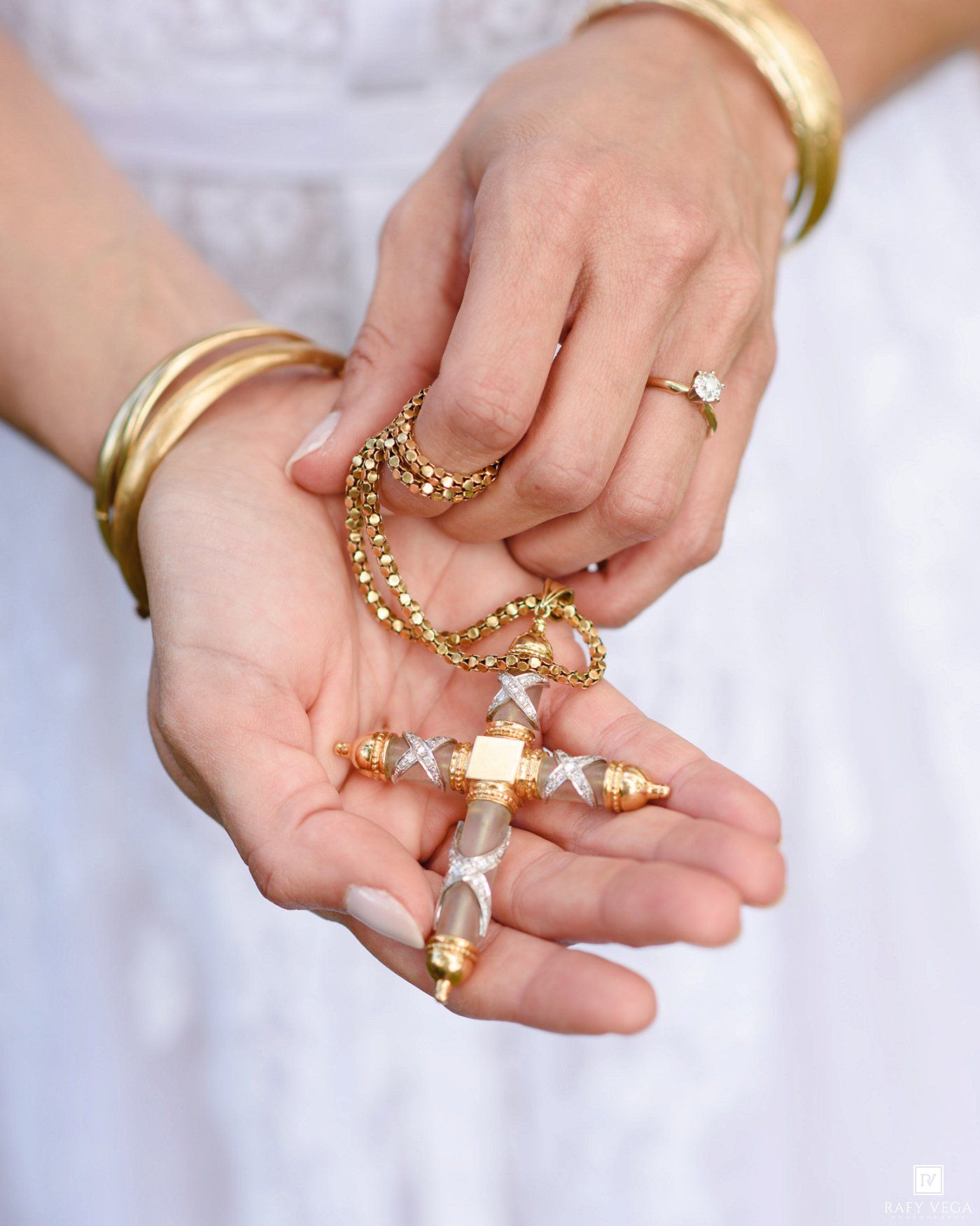 Hands holding a gold chain necklace with a crystal cross pendant, wearing gold bangles and a diamond ring.