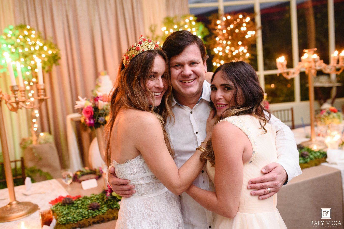 Two brides in white dresses and a man smile together at an elegant wedding reception with floral decor.