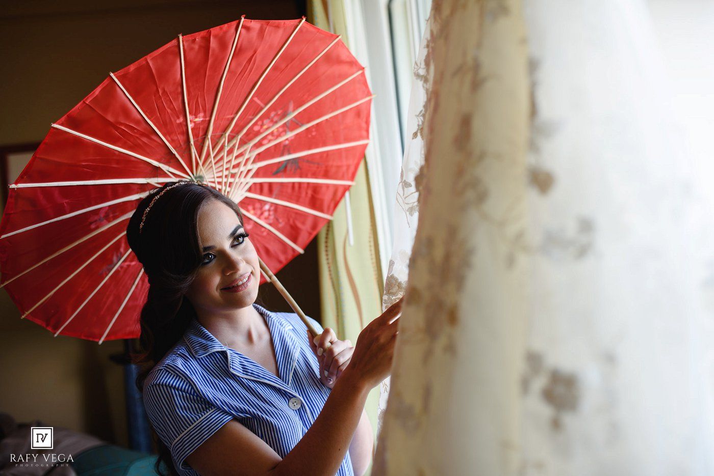 A smiling bride holds a red parasol umbrella near a window, wearing a blue dress during wedding preparations.