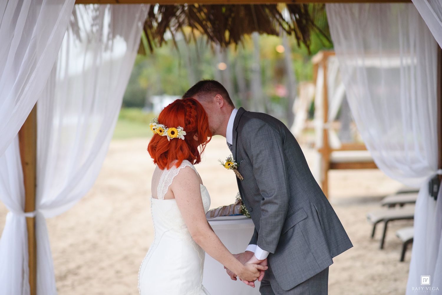 Bride and groom share first kiss under white draped wedding arch on sandy beach surrounded by tropical trees.