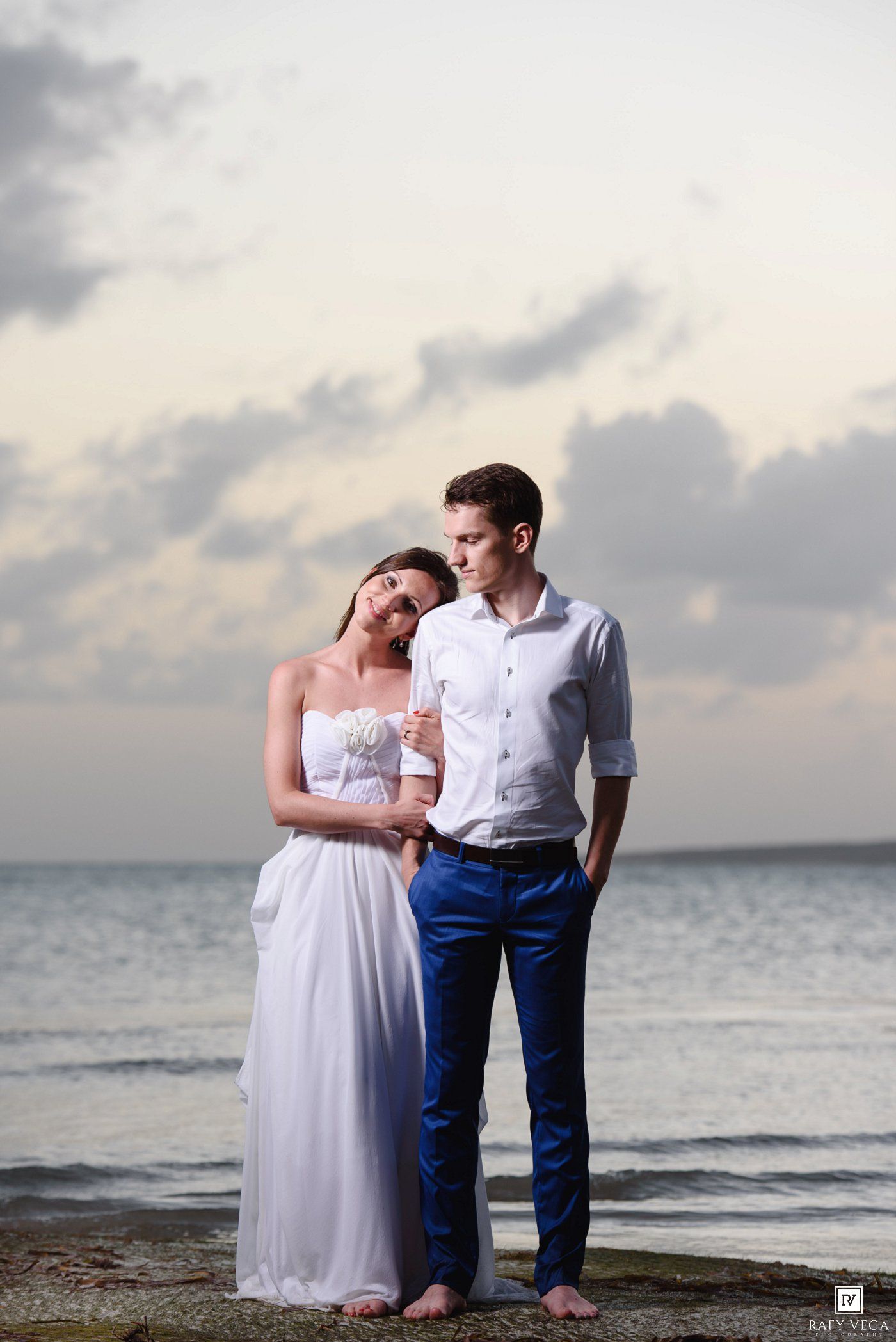 A couple in wedding attire stands barefoot on a rocky beach at dusk, the bride in white gown leaning on the groom.