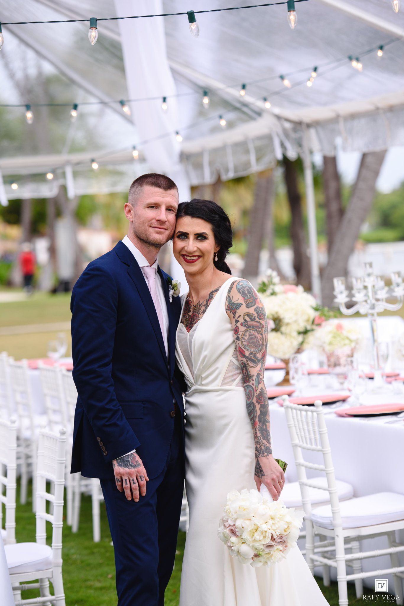 Bride with sleeve tattoos in white gown holds bouquet beside groom in navy suit at elegant outdoor wedding reception.