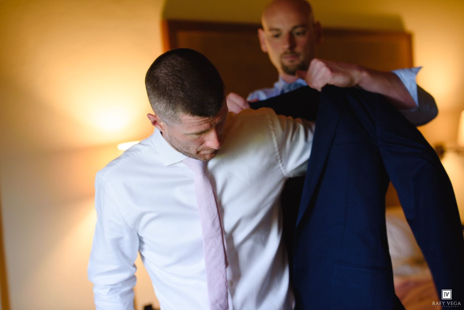 Groomsman helping groom put on navy suit jacket before wedding ceremony in warmly lit hotel room.