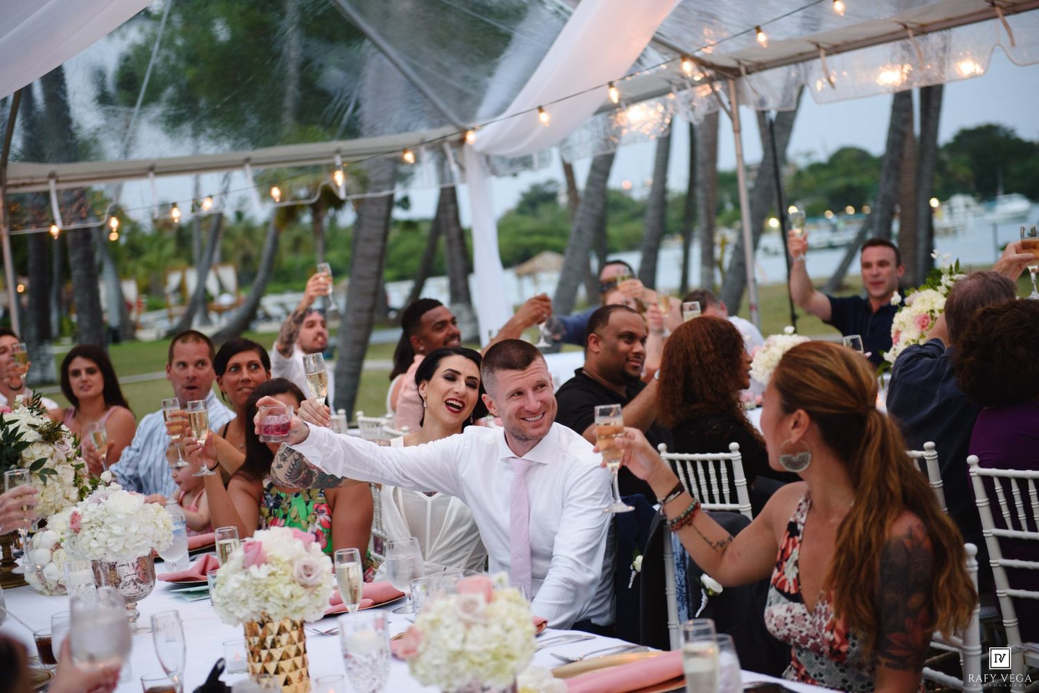 Guests raise glasses in a toast under a elegant outdoor wedding reception tent adorned with string lights and floral centerpieces.