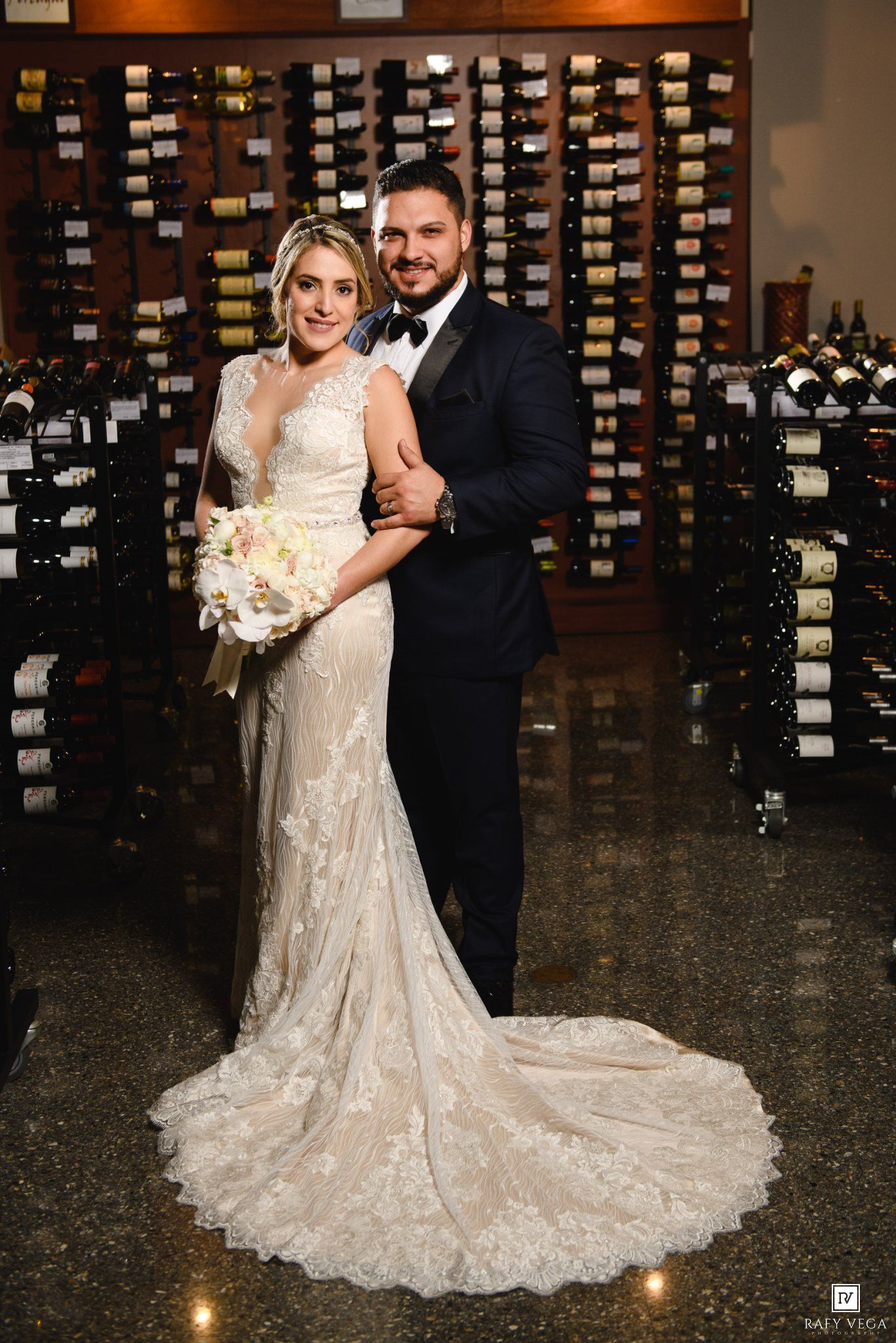 Bride and groom pose elegantly in a wine cellar, she in lace gown with bouquet, he in black tuxedo.