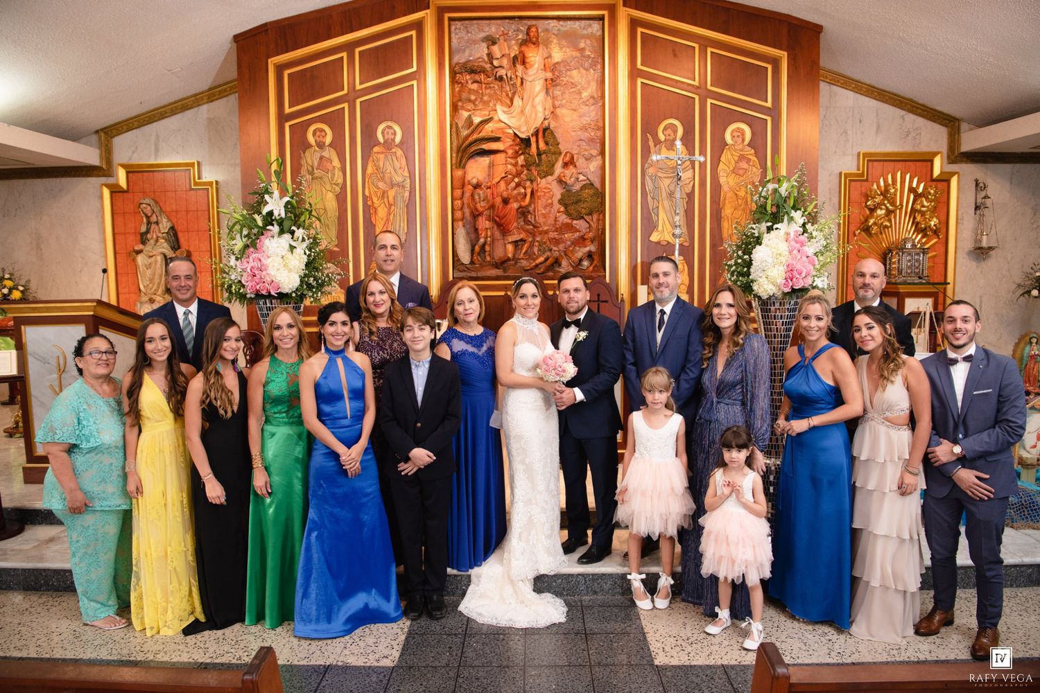 Wedding party poses together in a church with ornate altar, featuring bride and groom surrounded by family in colorful formal attire.