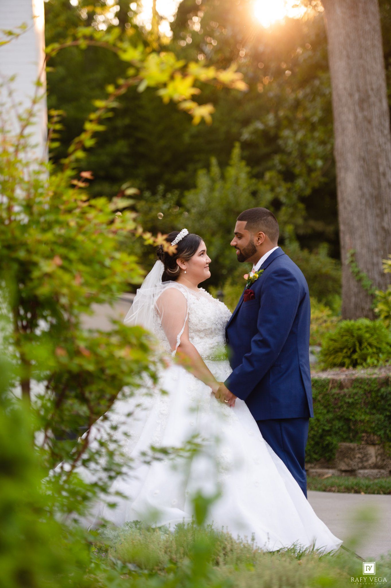 Bride and groom share a romantic moment in a lush garden at golden hour, she in white gown, he in navy suit.