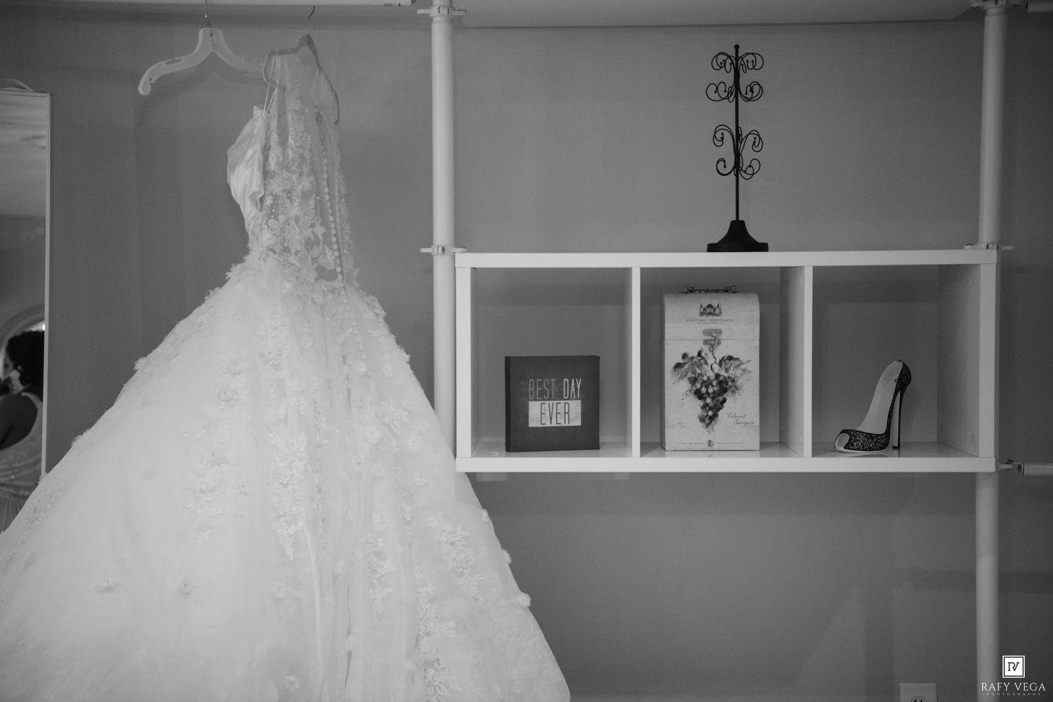 Black and white photo of an elegant wedding dress hanging near a white shelf with decorative items and a high heel shoe.
