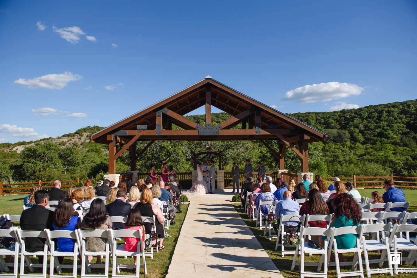 Outdoor wedding ceremony under a wooden pavilion with guests seated along an aisle surrounded by green hills.