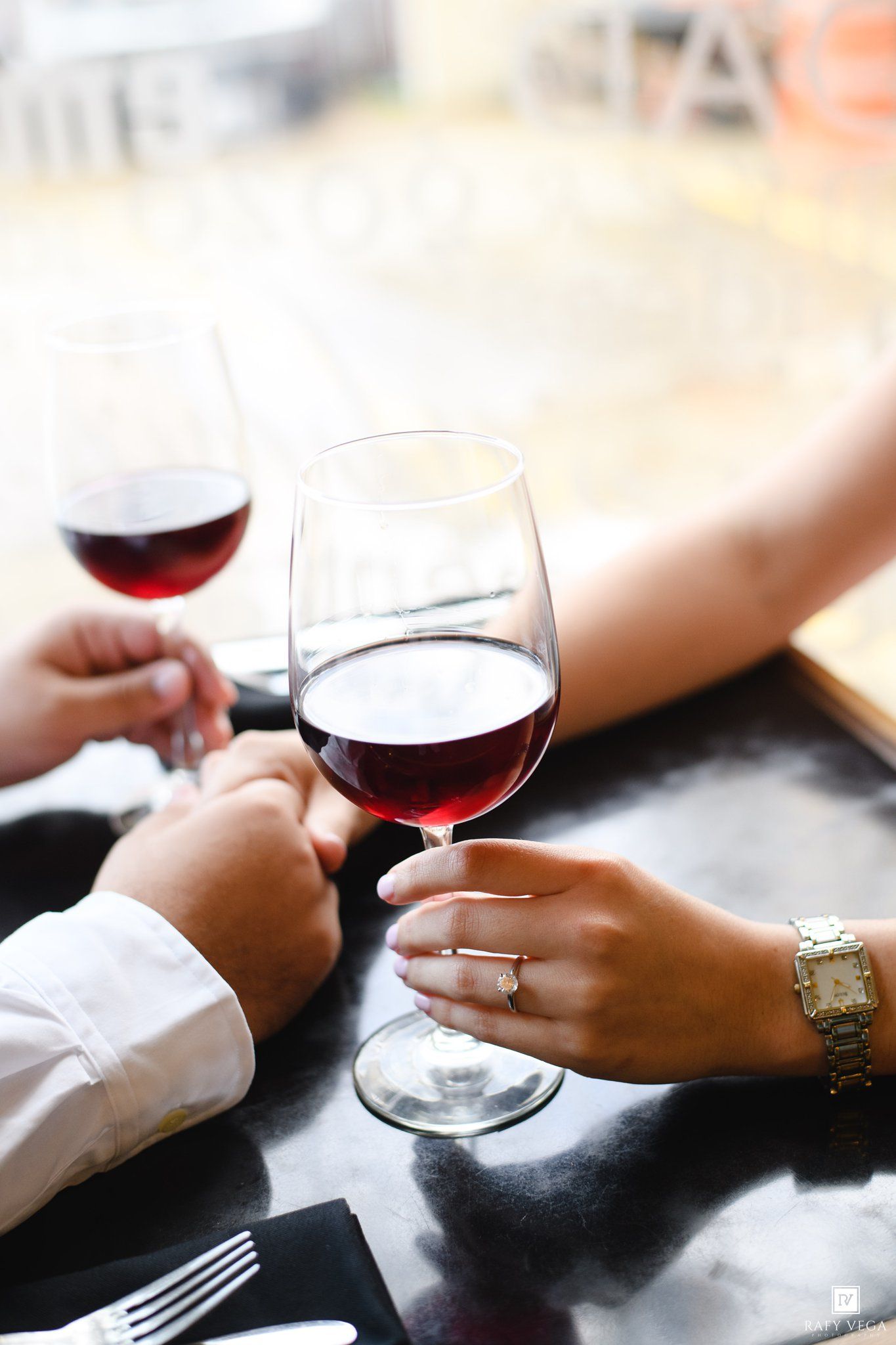Couple toasting with red wine glasses at a restaurant, woman wearing diamond ring and gold watch.