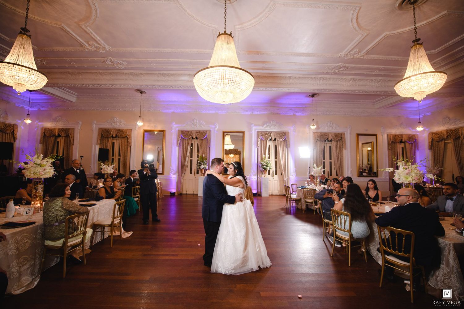Bride and groom share first dance in elegant ballroom with chandeliers and guests seated at decorated tables.