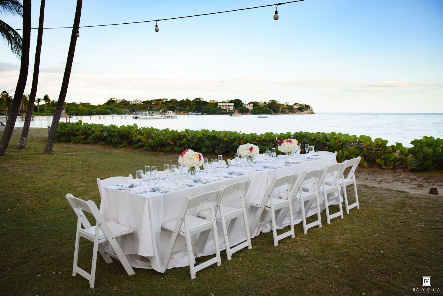 Elegant outdoor beachside wedding reception with long white table, chairs, and floral centerpieces at sunset.