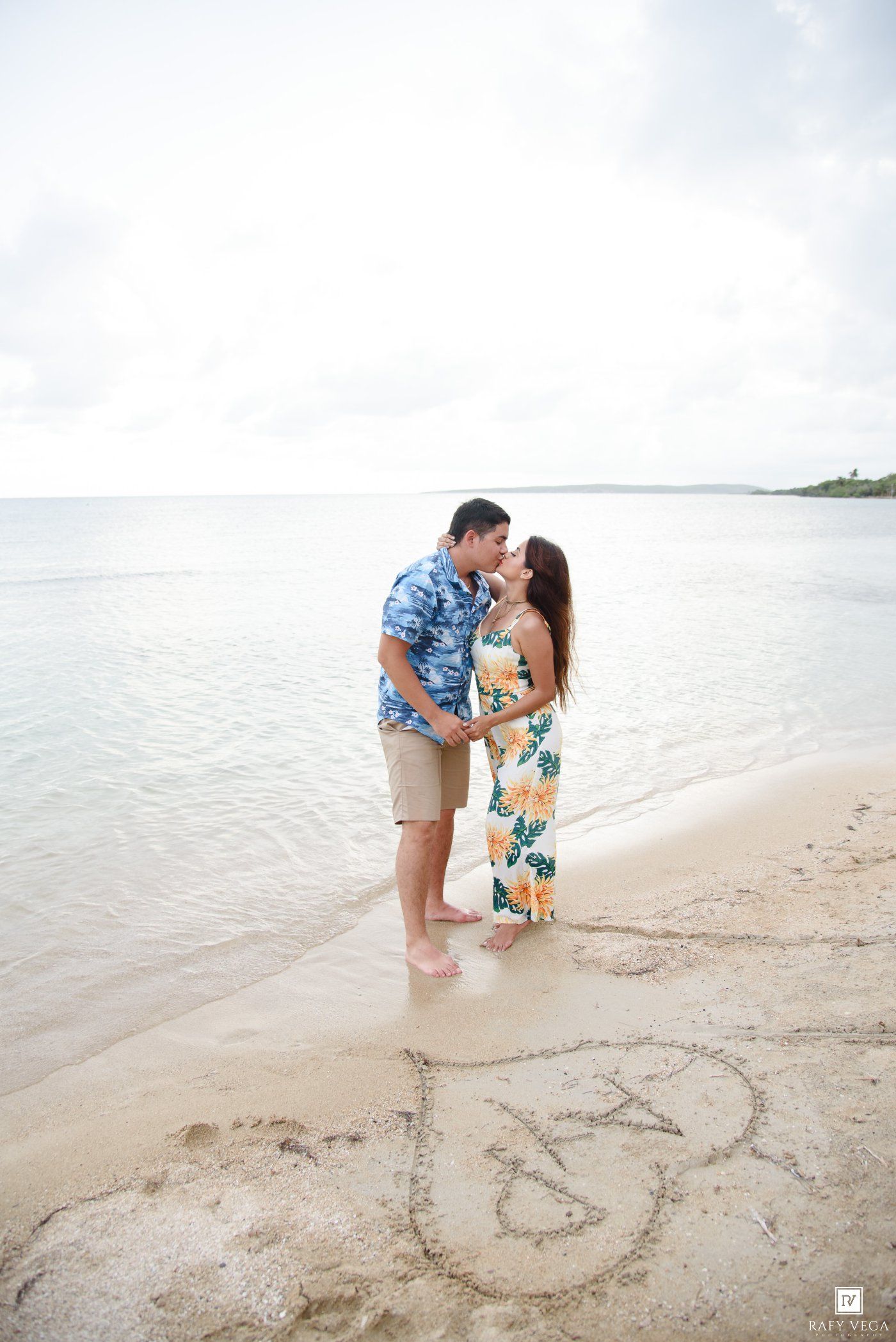 Couple kissing on a tropical beach with hearts drawn in the sand, wearing floral outfits near calm ocean waters.