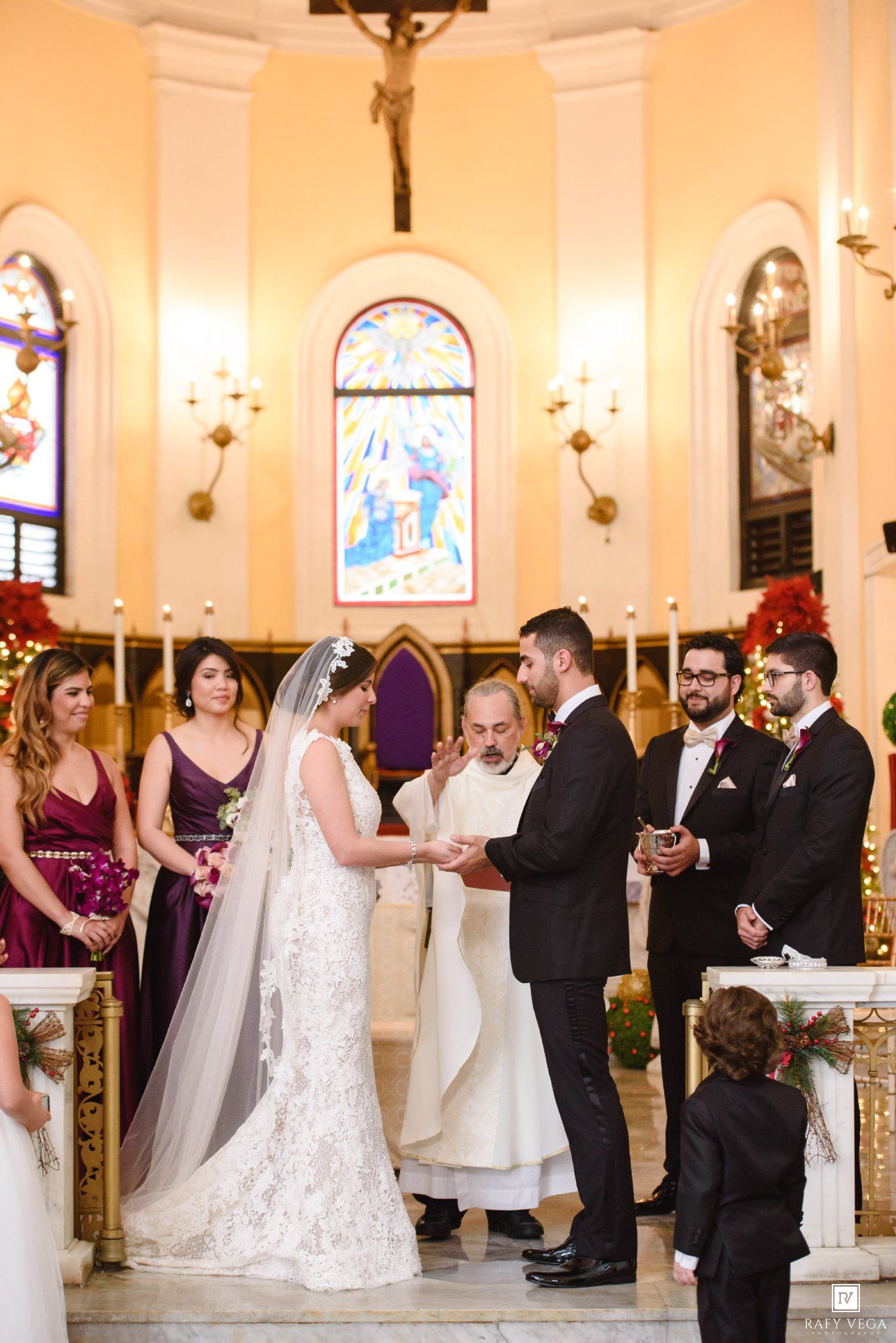 Bride and groom exchange vows at elegant church altar with priest, bridesmaids in burgundy, and stained glass windows.