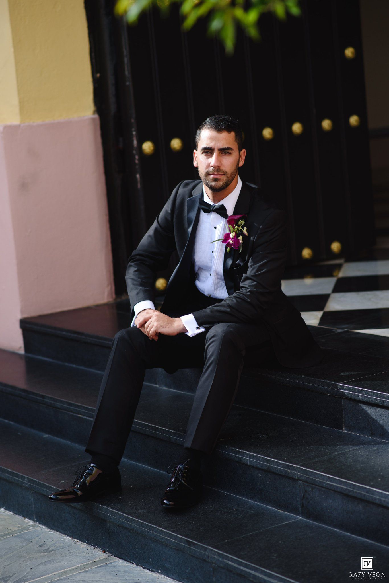 Groom in black tuxedo with boutonniere sitting on steps before wedding ceremony.