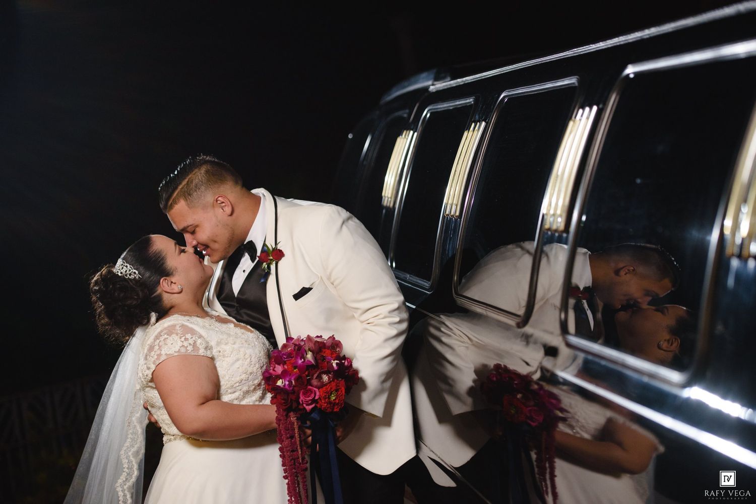 Bride and groom sharing a romantic kiss beside a black limousine at night during their wedding celebration.