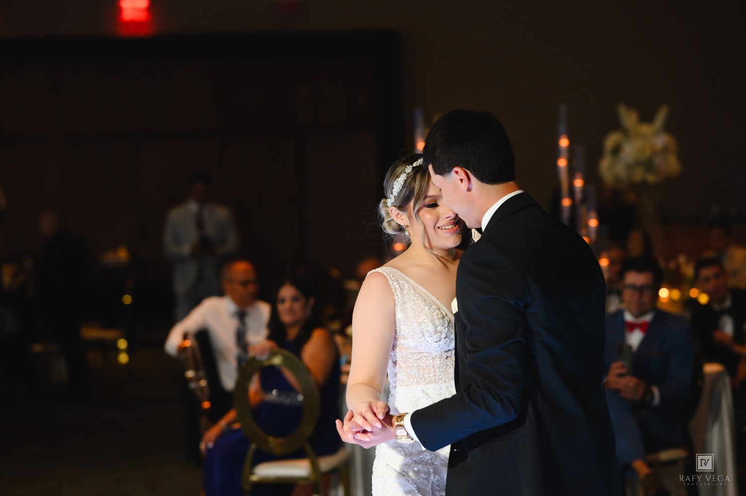 Bride and groom share their first dance at a wedding reception, smiling romantically under warm venue lighting.