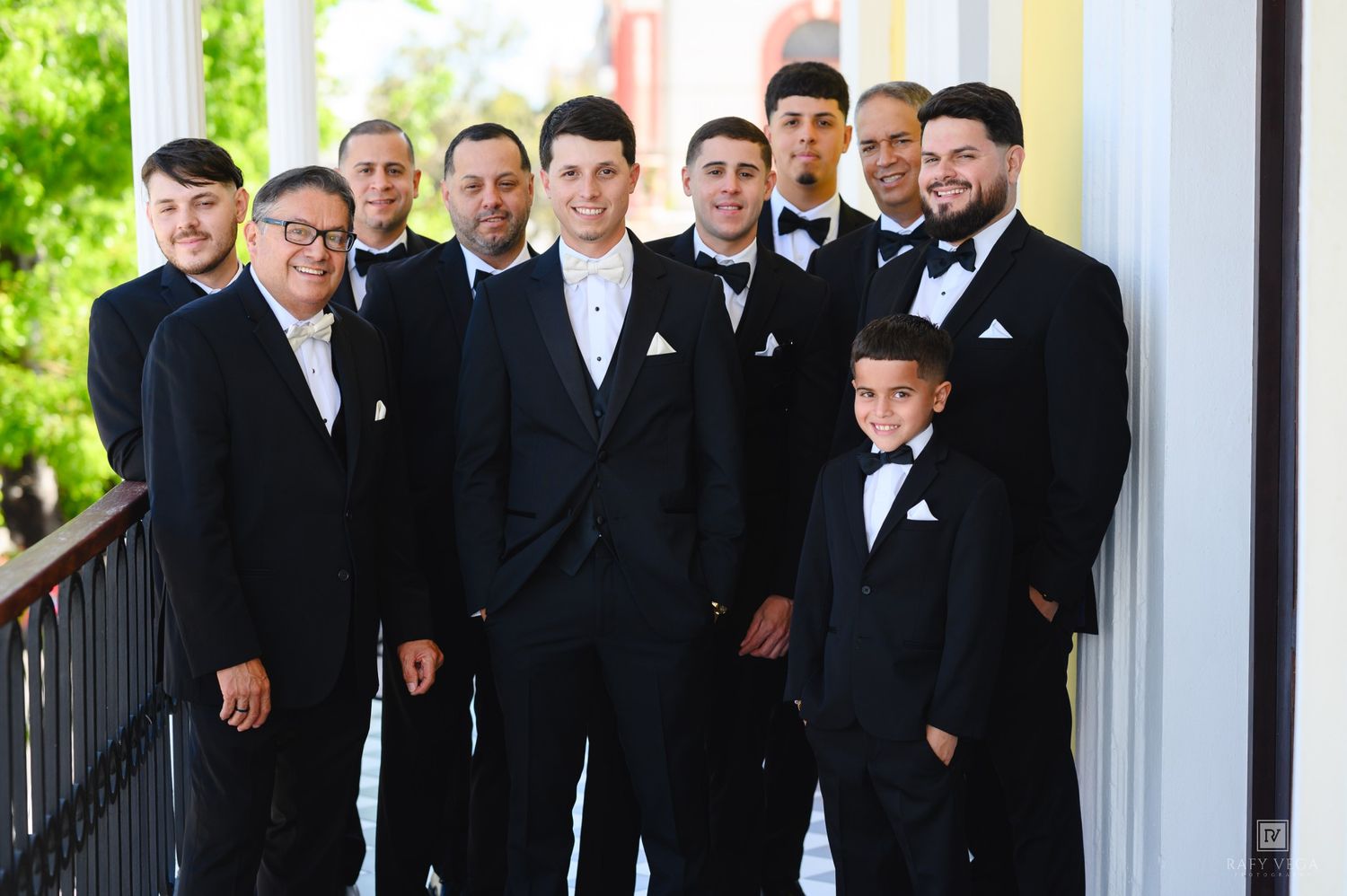 Groom and groomsmen in black tuxedos with bow ties posing on a balcony at a wedding.