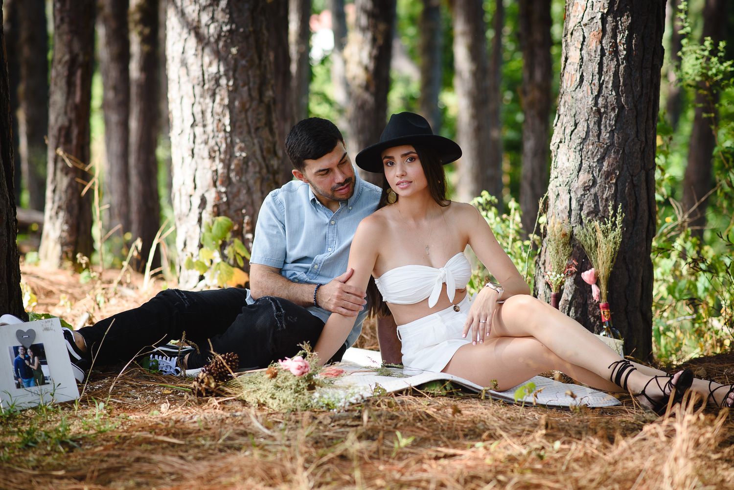 Couple relaxing on a picnic blanket in a pine forest, woman in white dress and black hat, man in blue shirt.