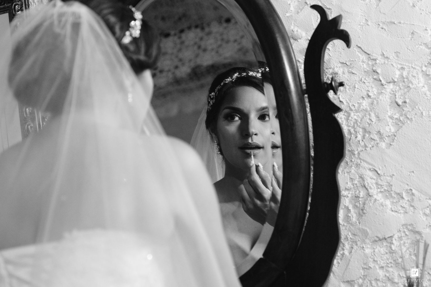 Black and white portrait of a bride in veil adjusting makeup while gazing into an ornate vintage mirror.