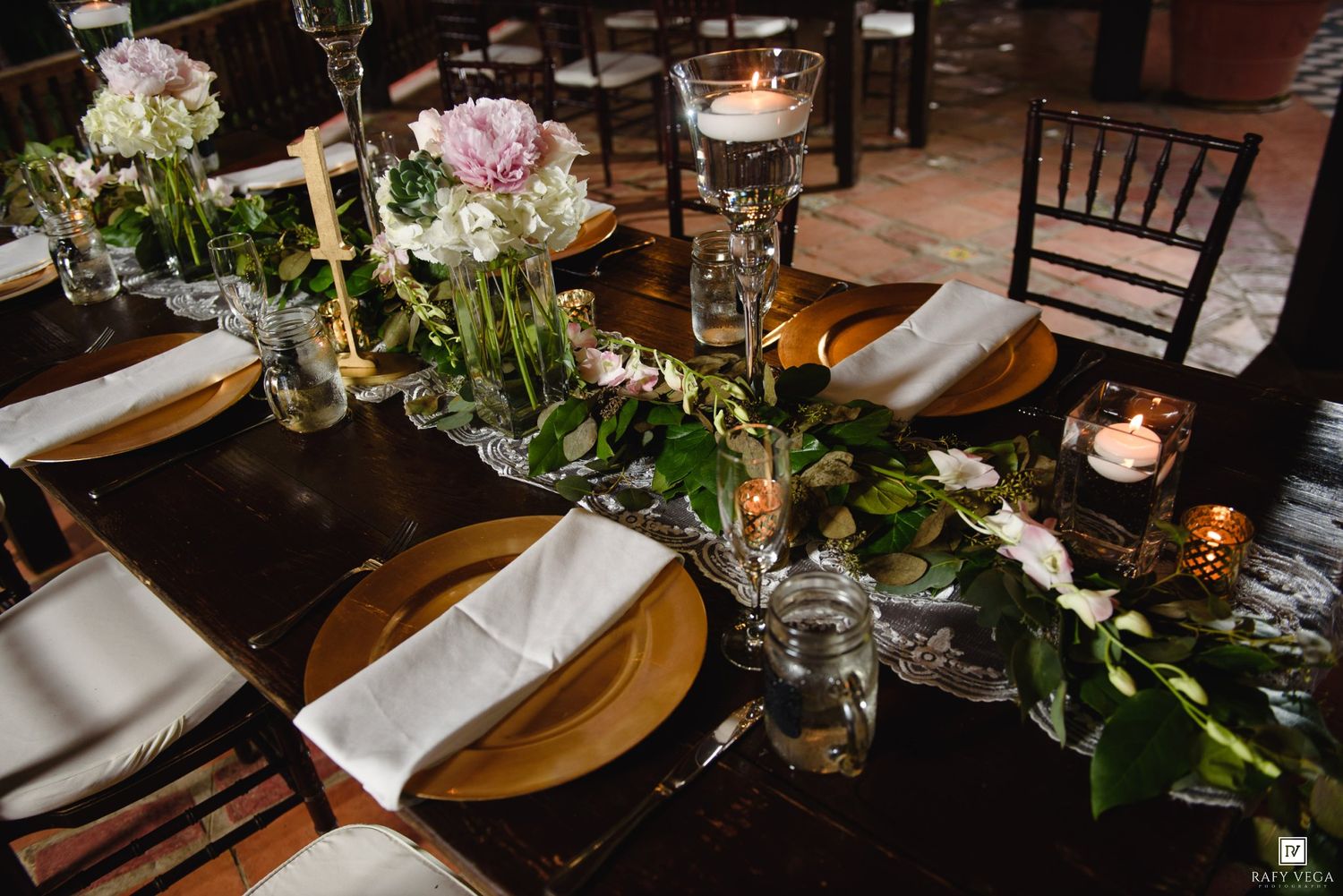 Elegant wedding reception table with gold chargers, white napkins, floral greenery runner, and glowing candles in a dark venue.