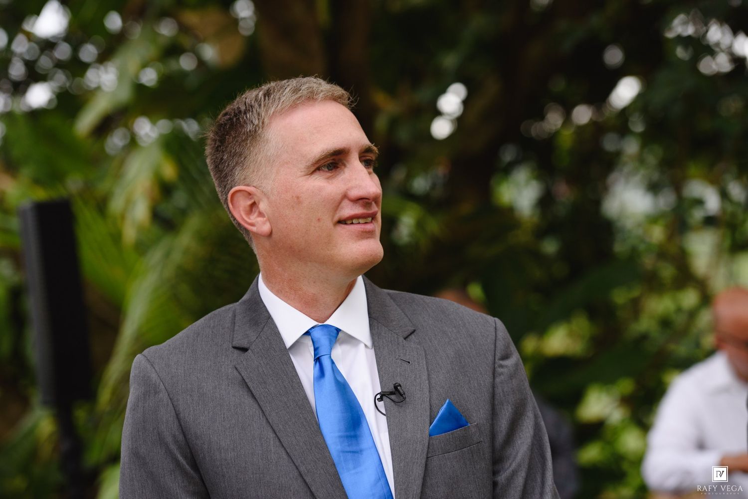Man in gray suit and blue tie smiling outdoors at a wedding ceremony with tropical greenery background.