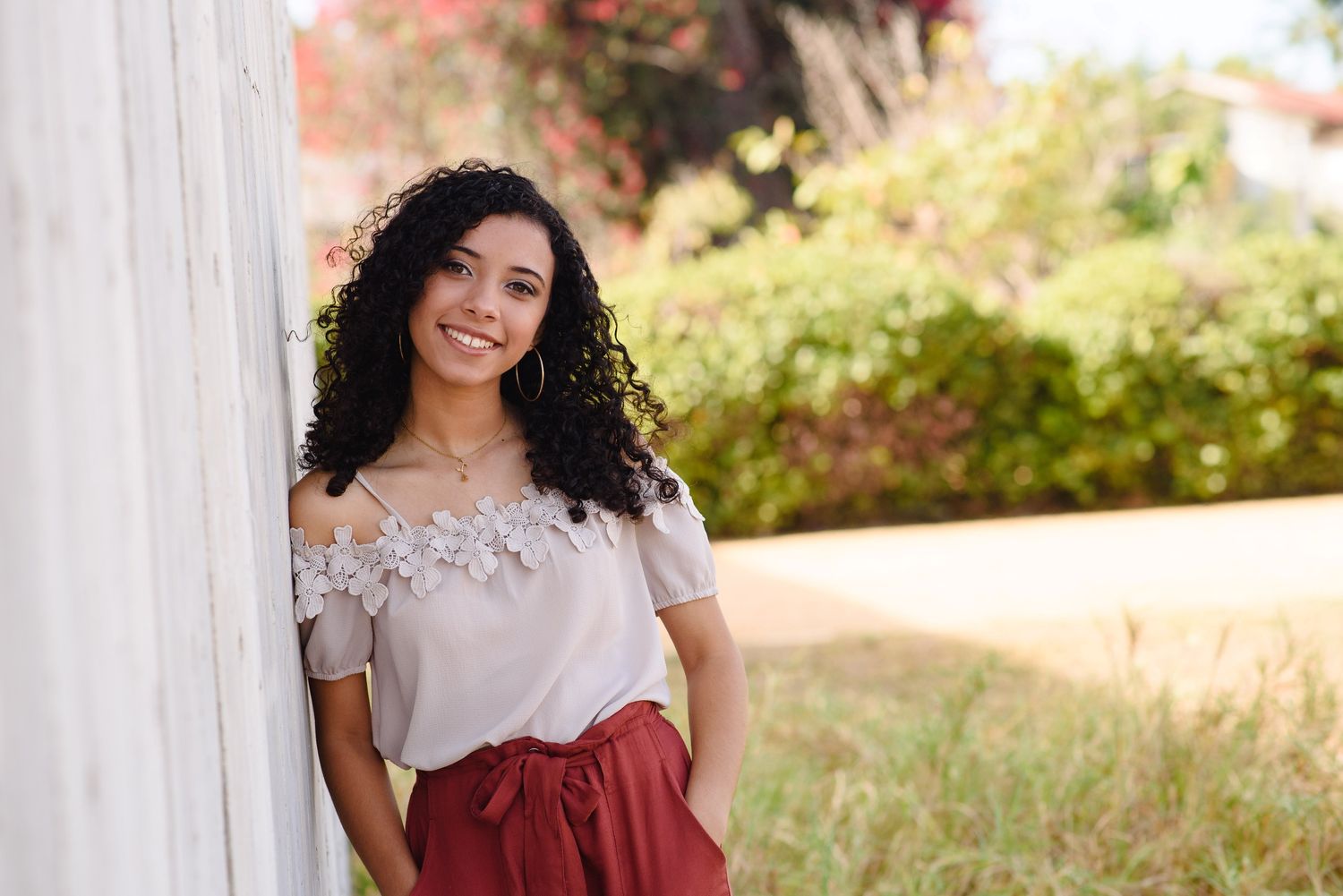A young woman with curly hair smiles while leaning against a white pillar outdoors in a floral top and red skirt.