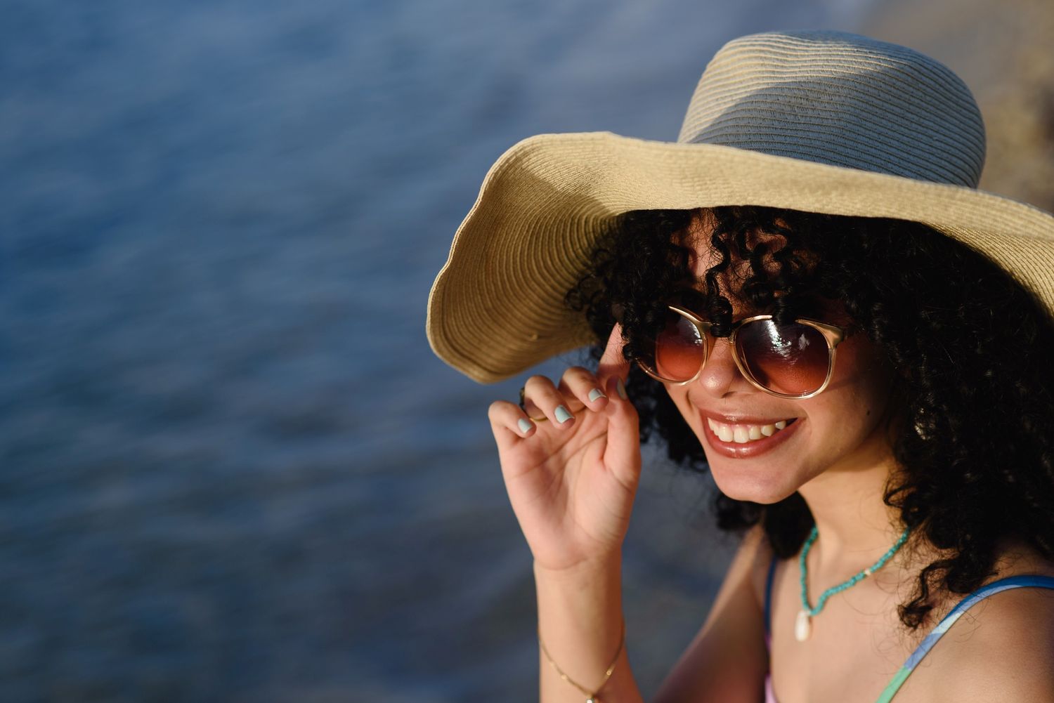 Smiling woman wearing a wide-brim straw hat and sunglasses near the ocean on a sunny day.