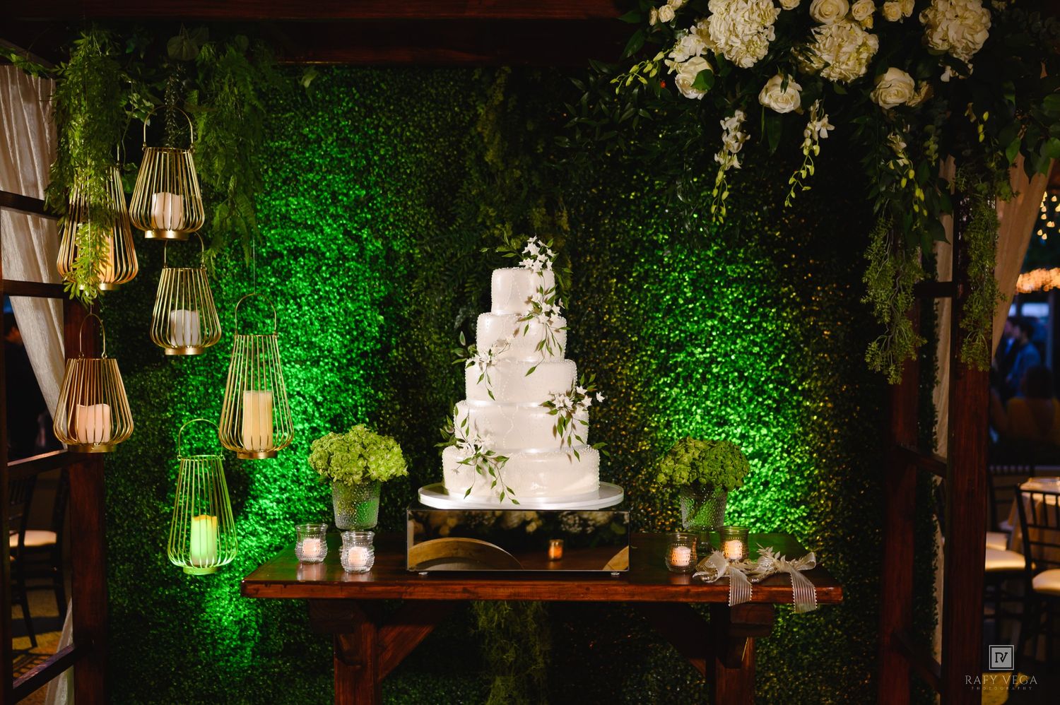 Elegant five-tier white wedding cake displayed on a wooden table against a lush green ivy wall with floral accents.