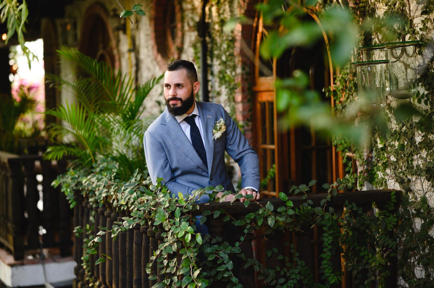 Groom in gray suit with boutonniere stands on vine-covered balcony surrounded by lush greenery.