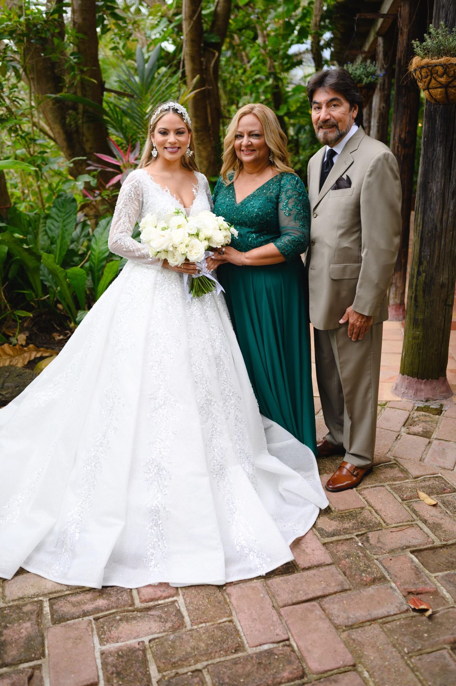 Bride in white lace gown holding bouquet poses with woman in green dress and man in tan suit at outdoor wedding.