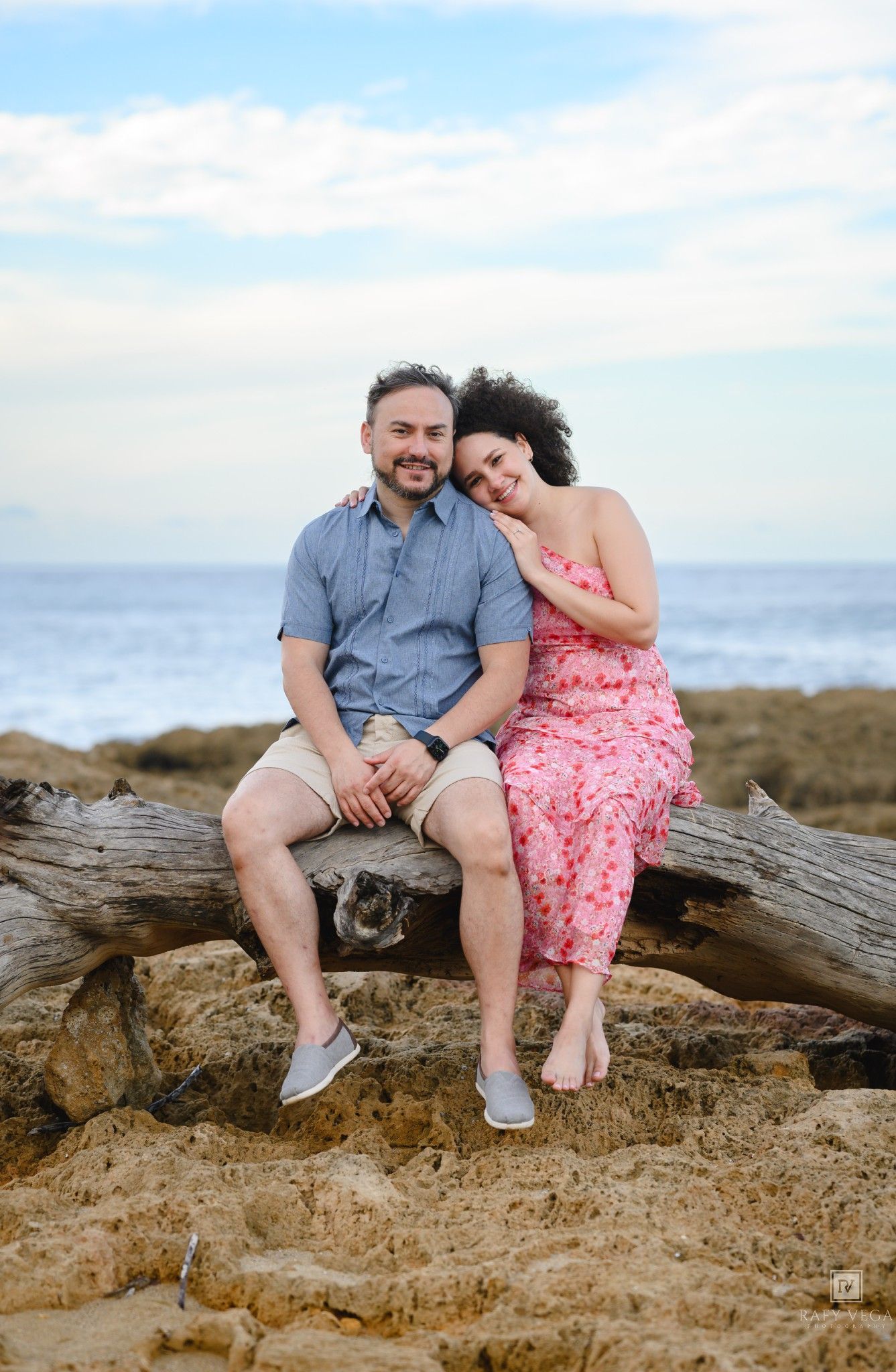 Couple sitting on driftwood log at beach, woman in pink floral dress leaning on man in blue shirt, ocean backdrop.