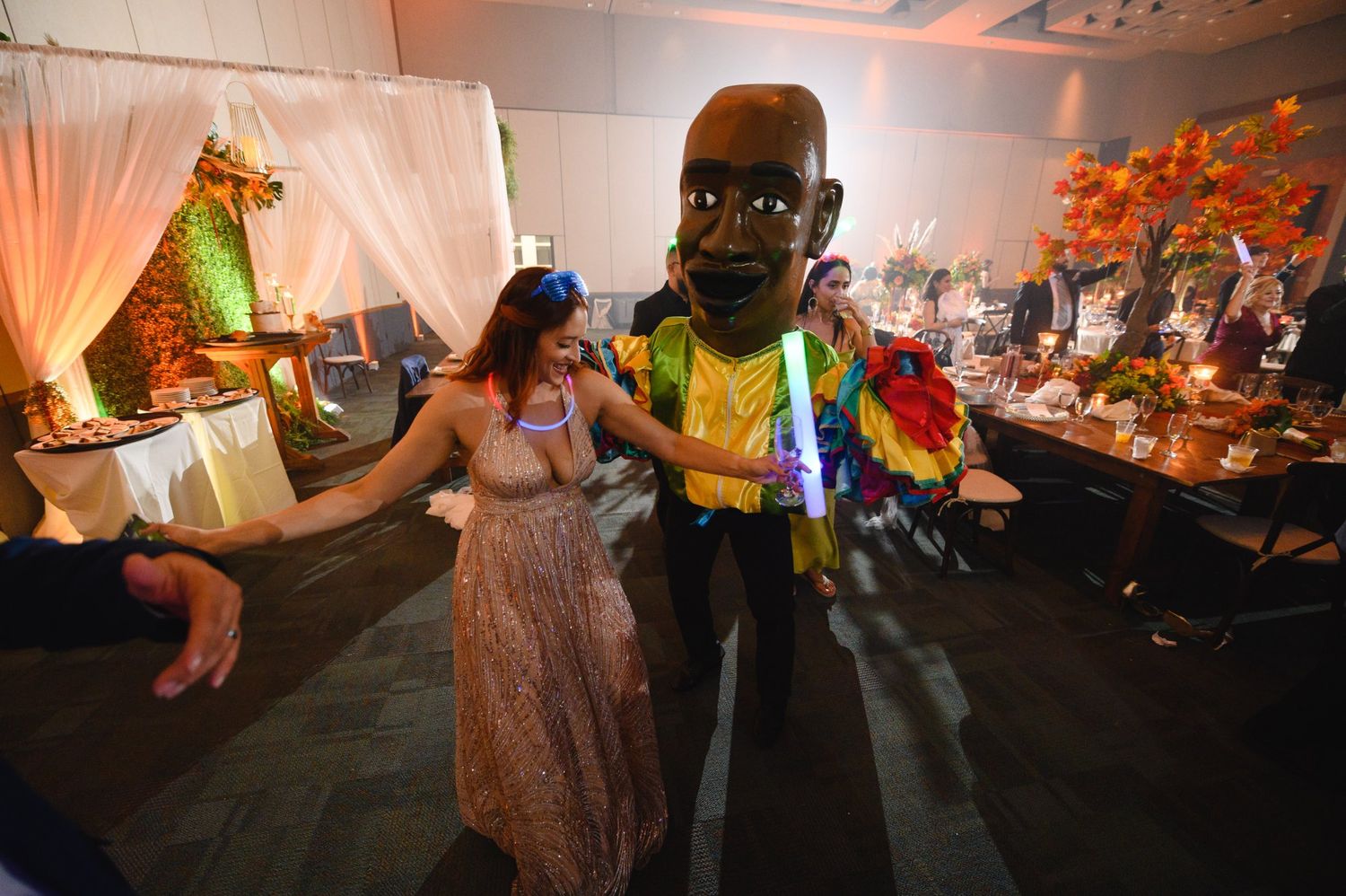 Woman in sequin dress dancing with a person in a giant cartoon head costume at a festive indoor event.