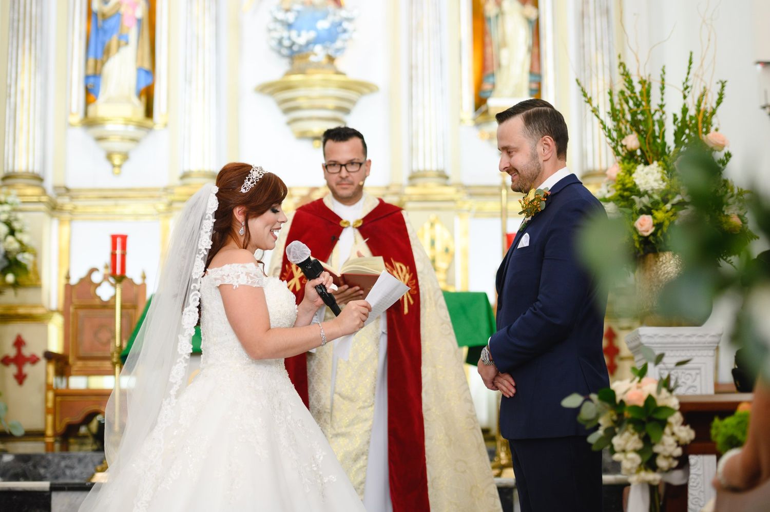 Bride and groom exchange vows at ornate church altar with priest in red vestments during wedding ceremony.