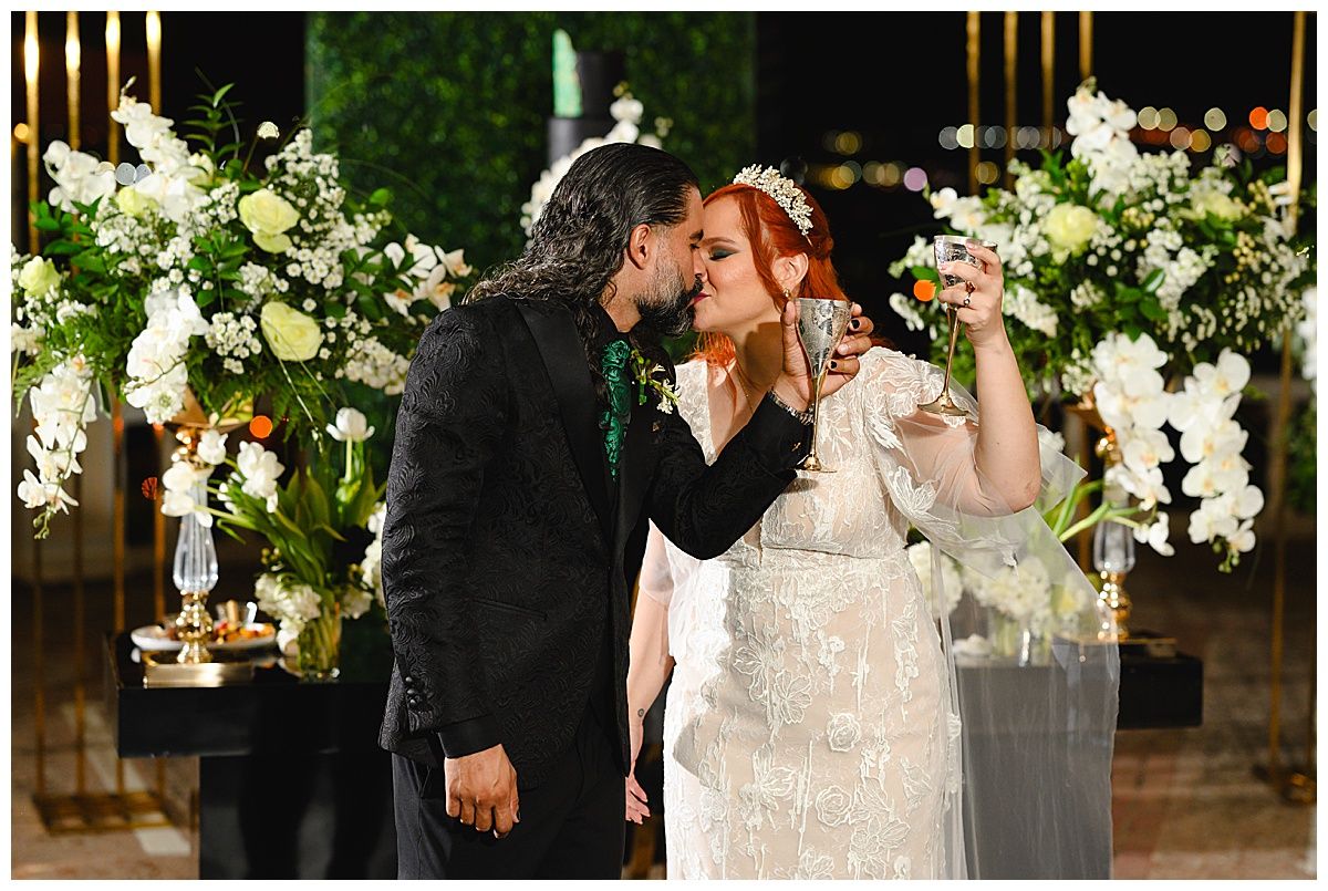 Bride and groom share a kiss while toasting champagne glasses at their elegant wedding reception with white floral arrangements.