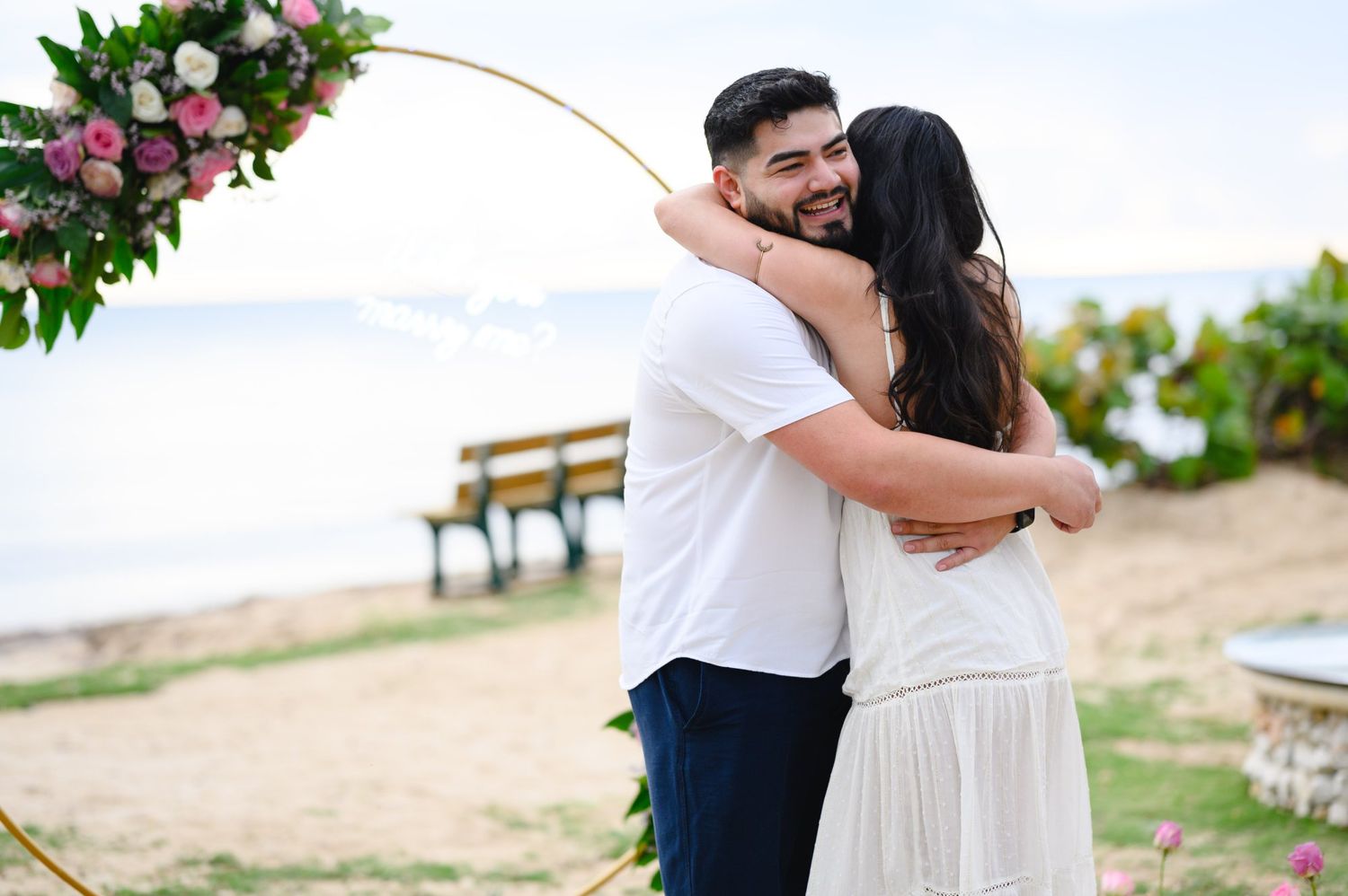 Couple embracing joyfully on a beach near a floral arch, celebrating a romantic proposal or wedding ceremony.