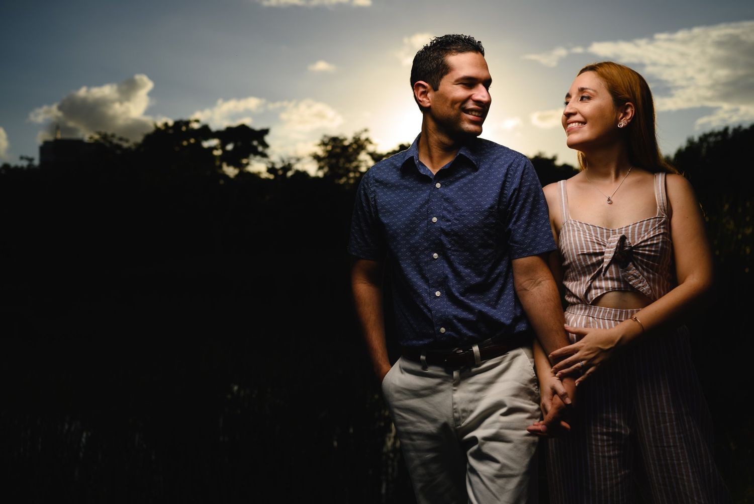 Couple holding hands smiling outdoors at sunset with dramatic sky and trees in background.