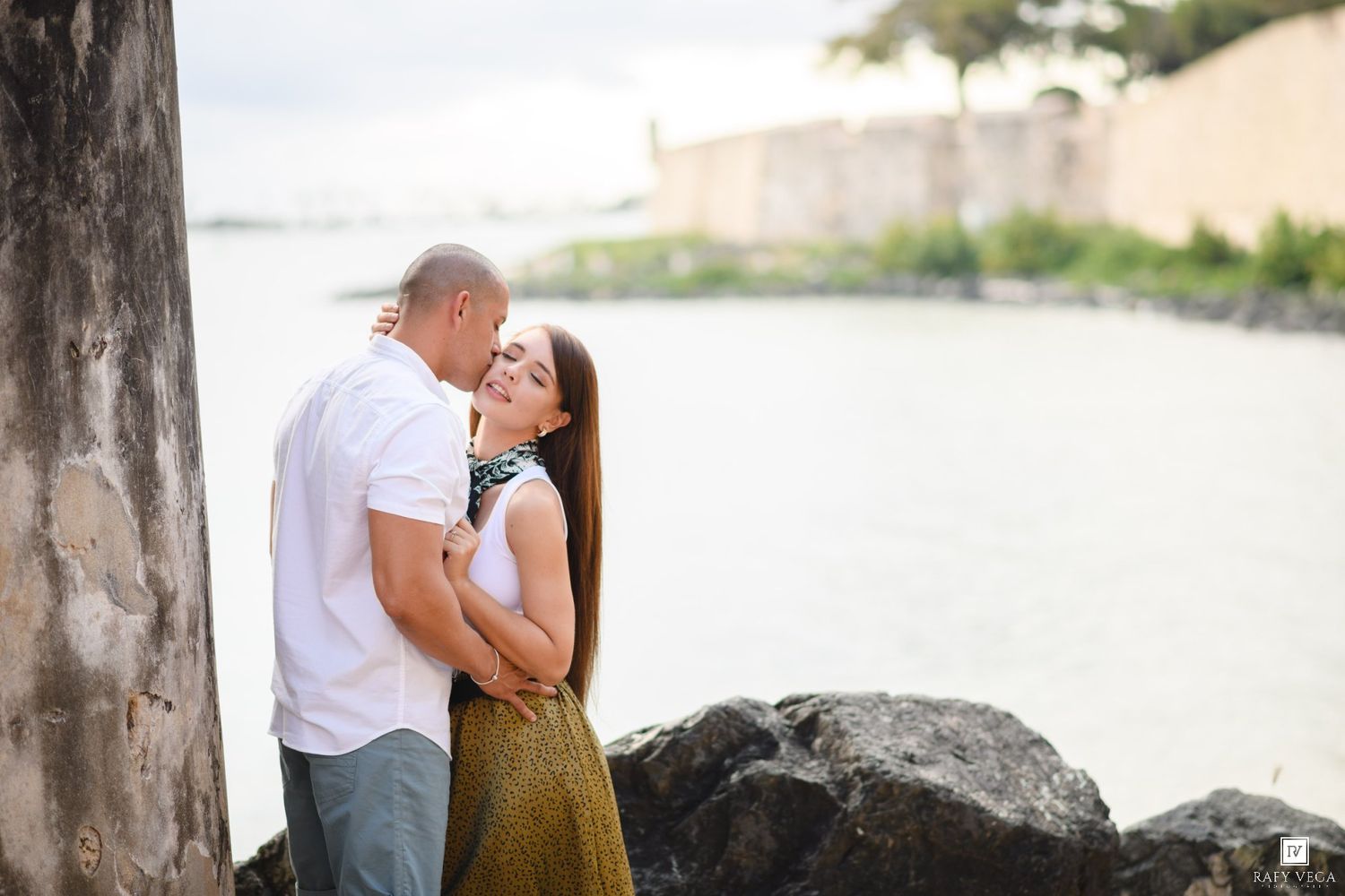 Couple embracing tenderly by rocky waterfront with historic stone walls and bay views behind them.