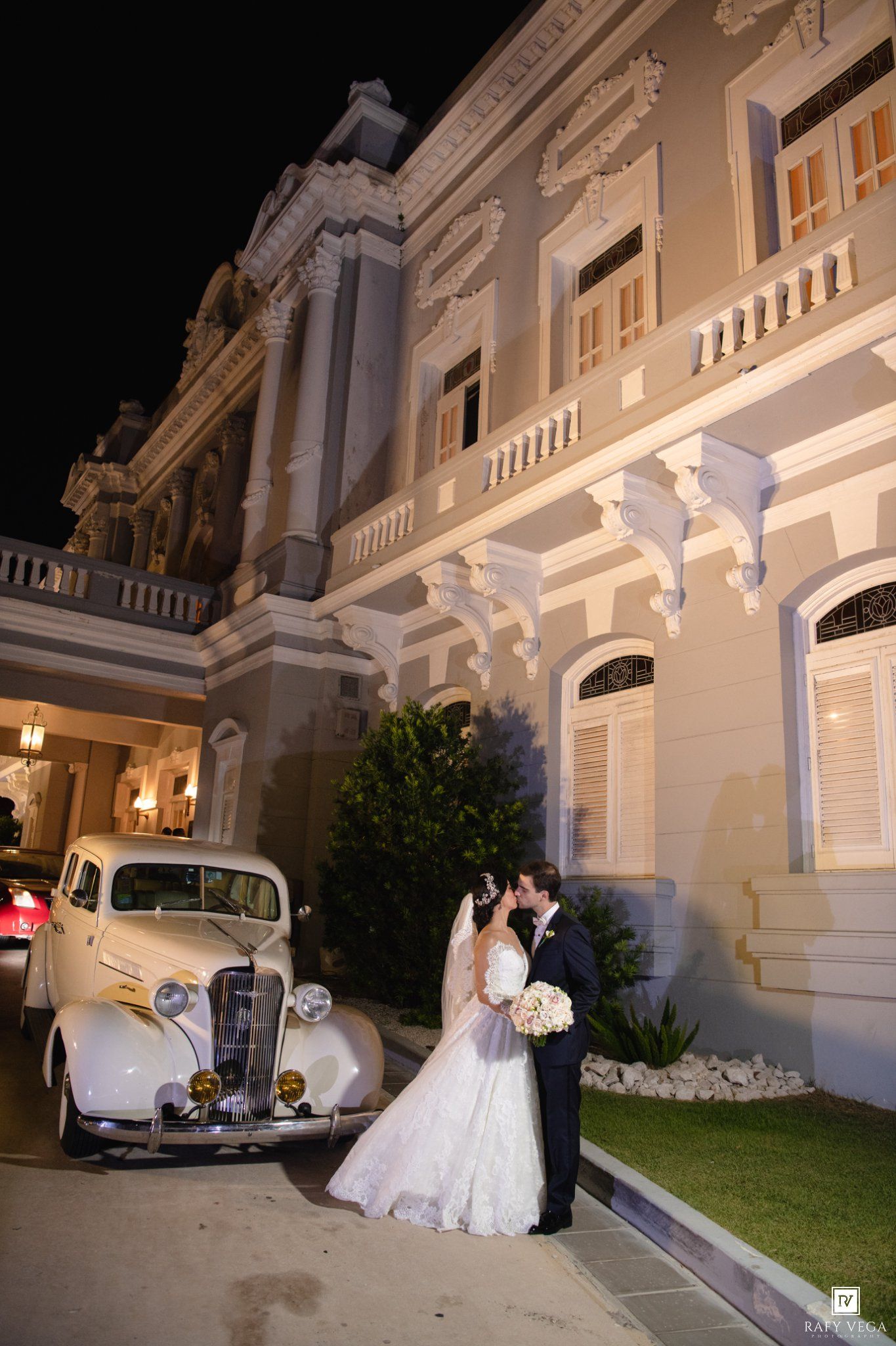 Bride and groom kissing beside a vintage white car outside an elegant illuminated mansion at night.