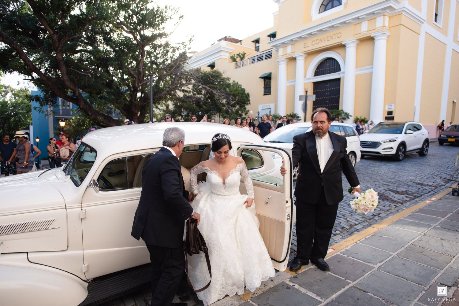 Bride in white gown exits vintage cream car assisted by two men outside a historic church on cobblestone street.